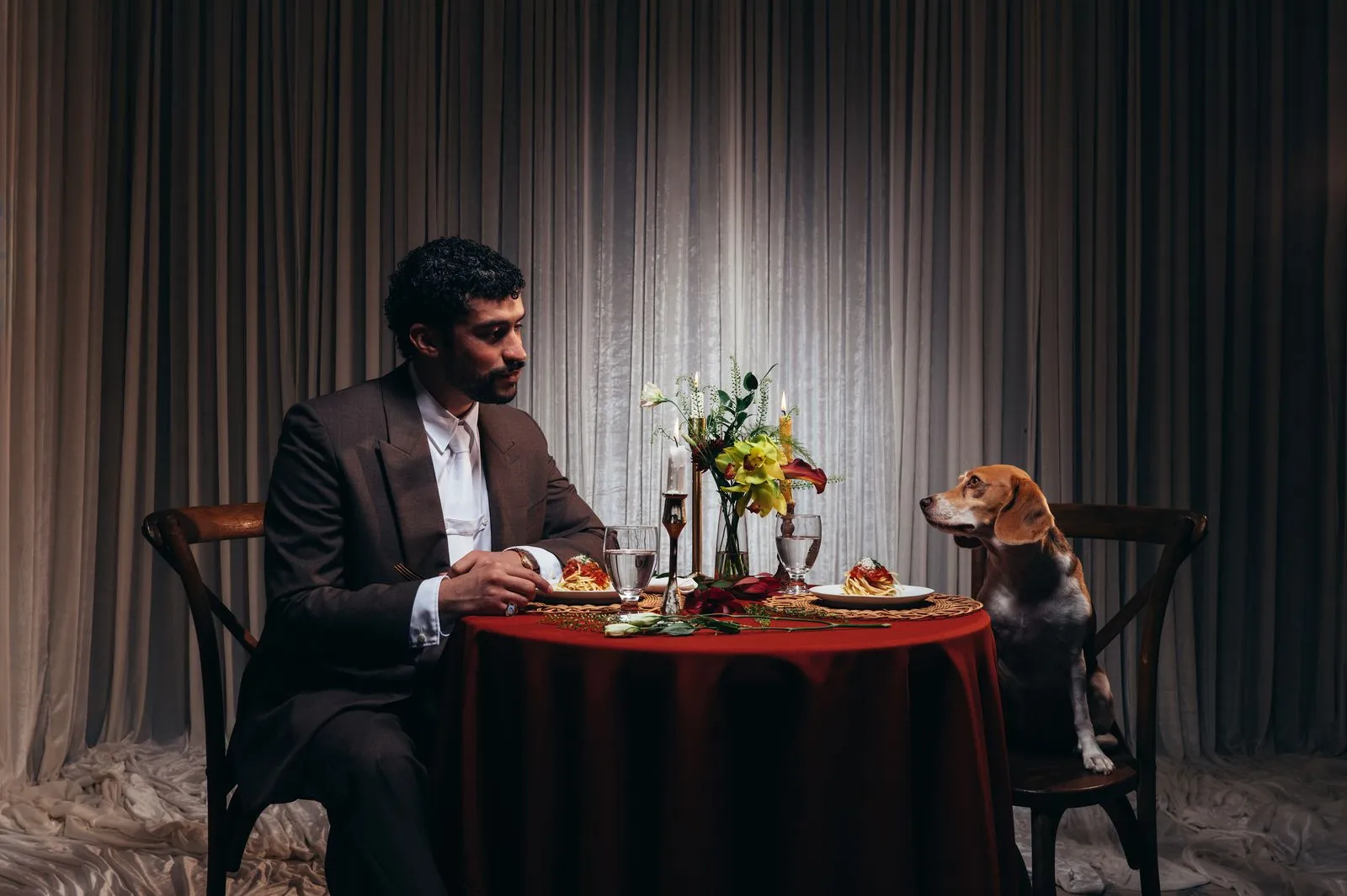 Man in a suit dining with a beagle at a candlelit table, pasta plate in front of each, floral centerpiece between them.
