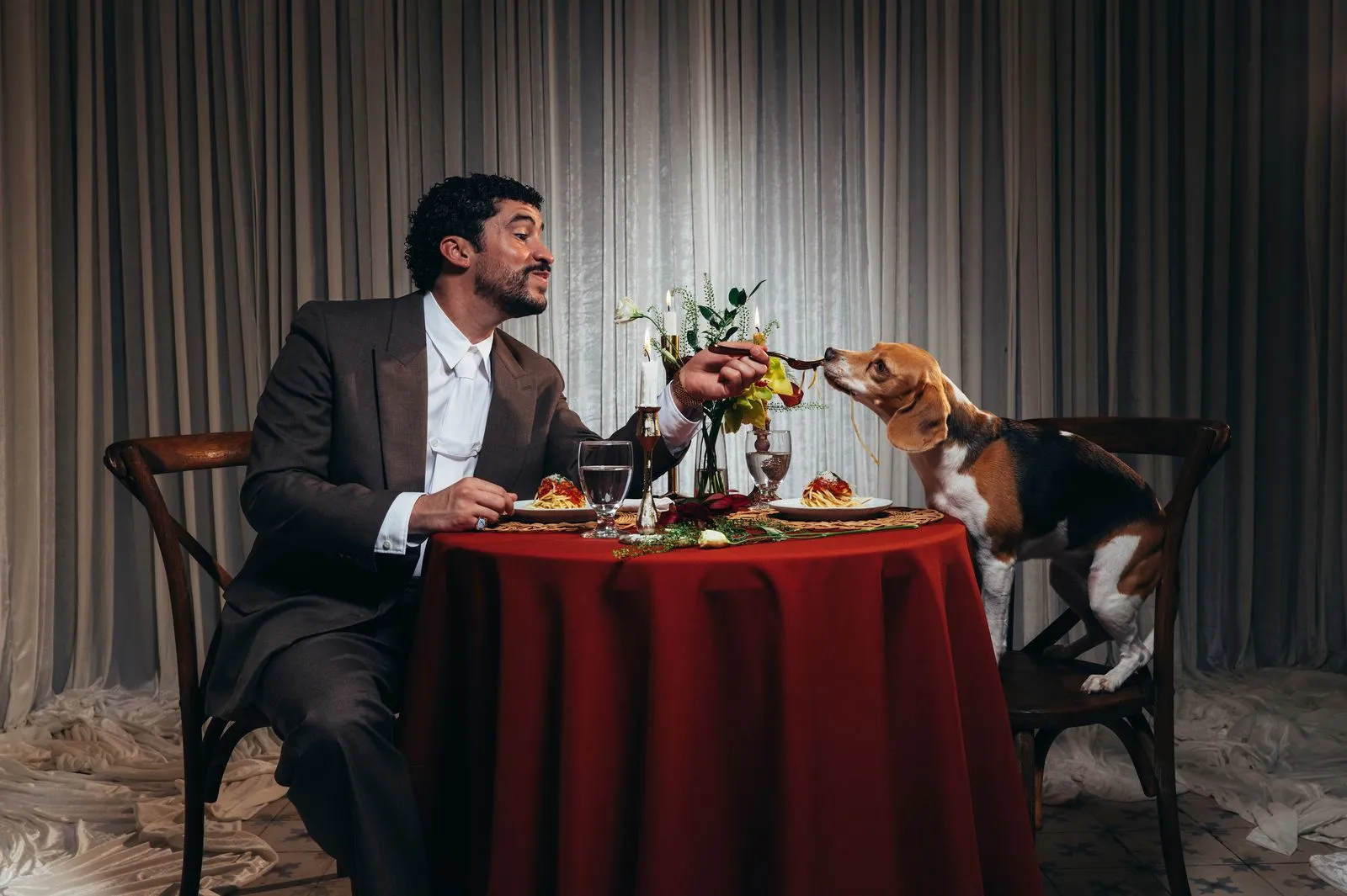Man in a suit shares a bite with a beagle at a formal dinner, passing a spoon across a round table with red tablecloth and flowers.