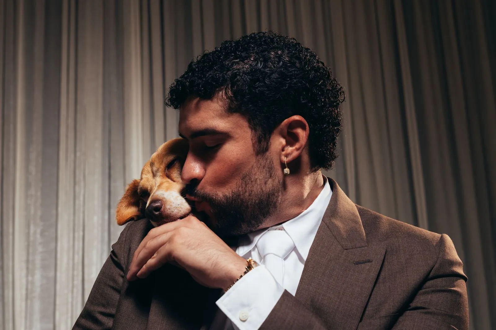 Man in a brown suit kisses a beagle on the nose, eyes closed in a tender moment behind a curtain backdrop.