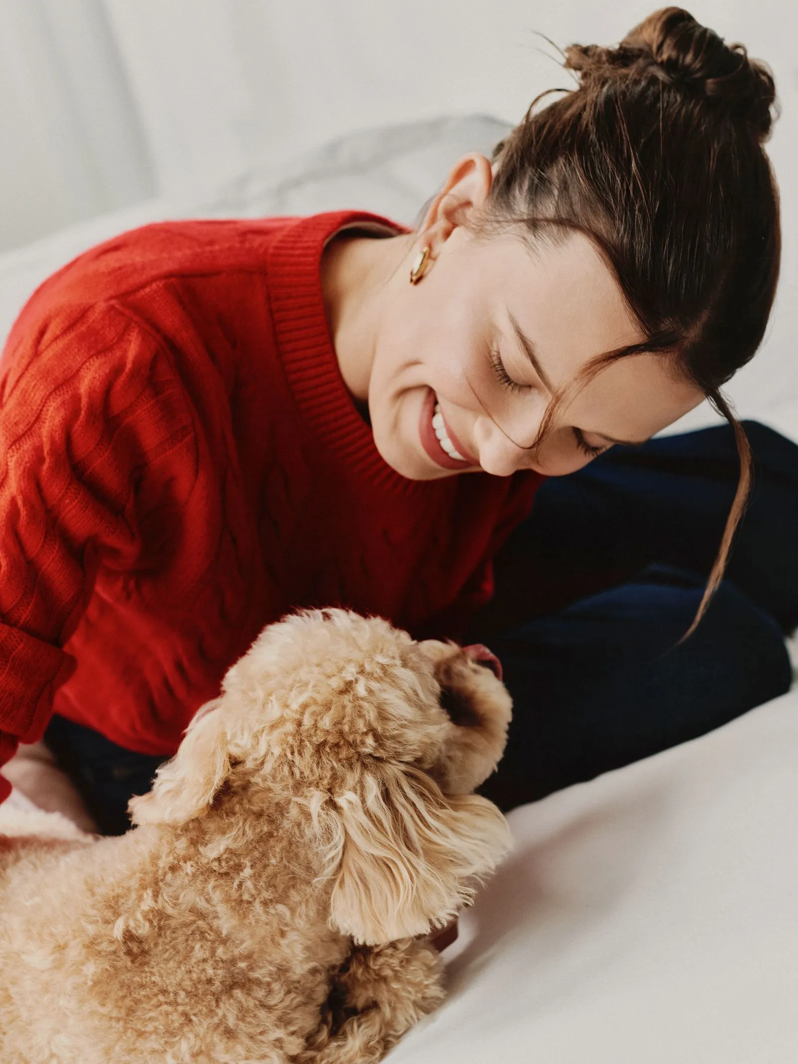 Woman in a red sweater smiling as she cuddles a fluffy beige dog on a bed.