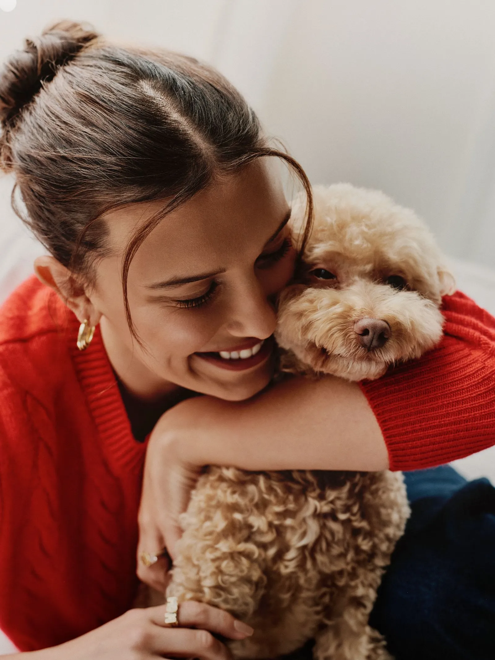 Woman in a red sweater cuddling a fluffy beige dog, smiling warmly at the camera (close embrace).
