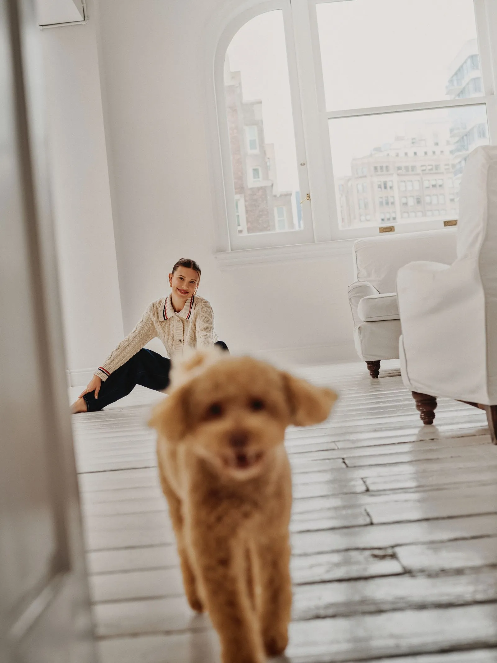 Woman sitting on a white wooden floor in a bright living room, with a small fluffy dog walking toward the camera.