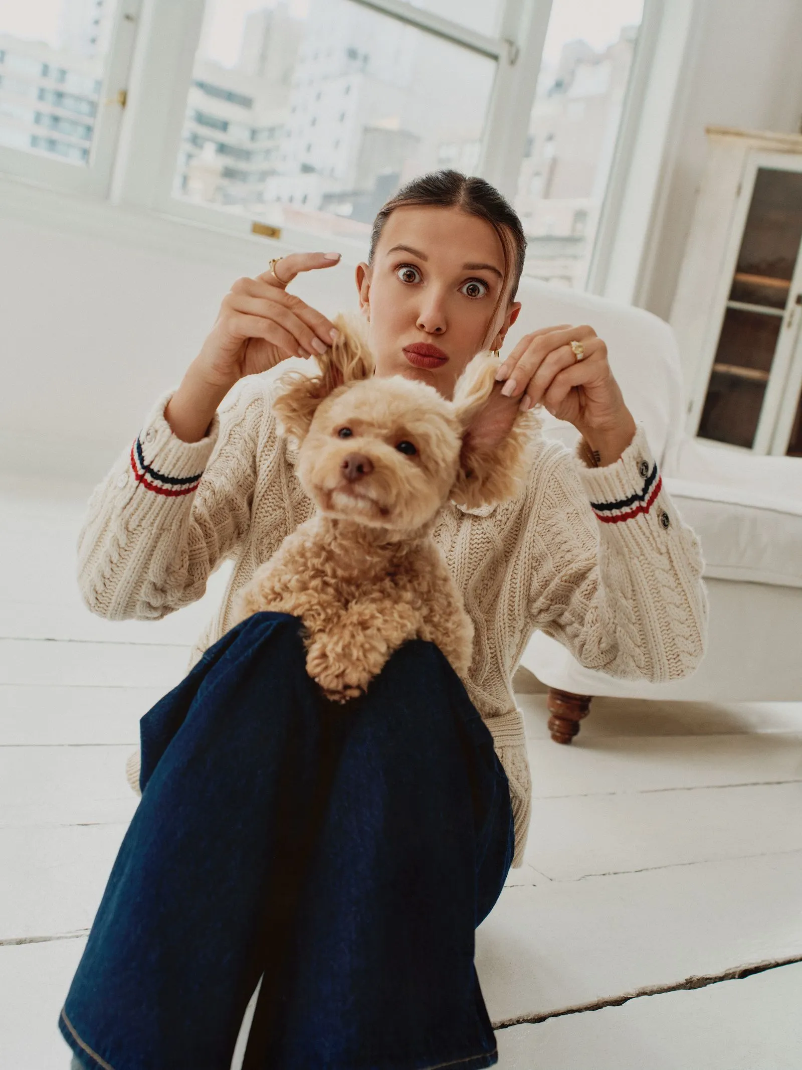 Woman in a cream knit sweater holds a fluffy beige dog toward the camera, indoors on a white wooden floor with a city view outside the windows.