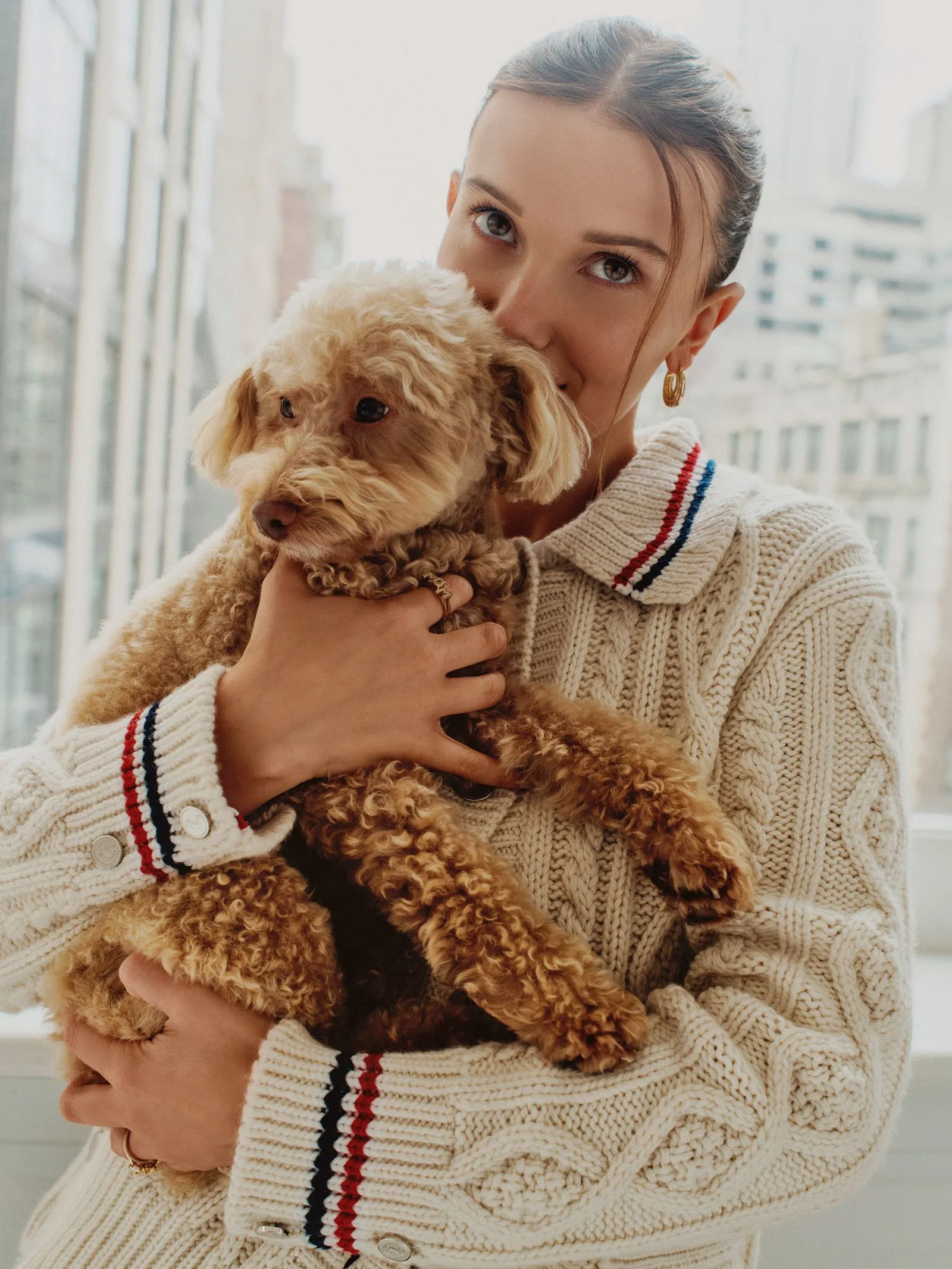 Woman in a cream knit sweater holding a fluffy brown poodle indoors by a window, looking at the camera and smiling.