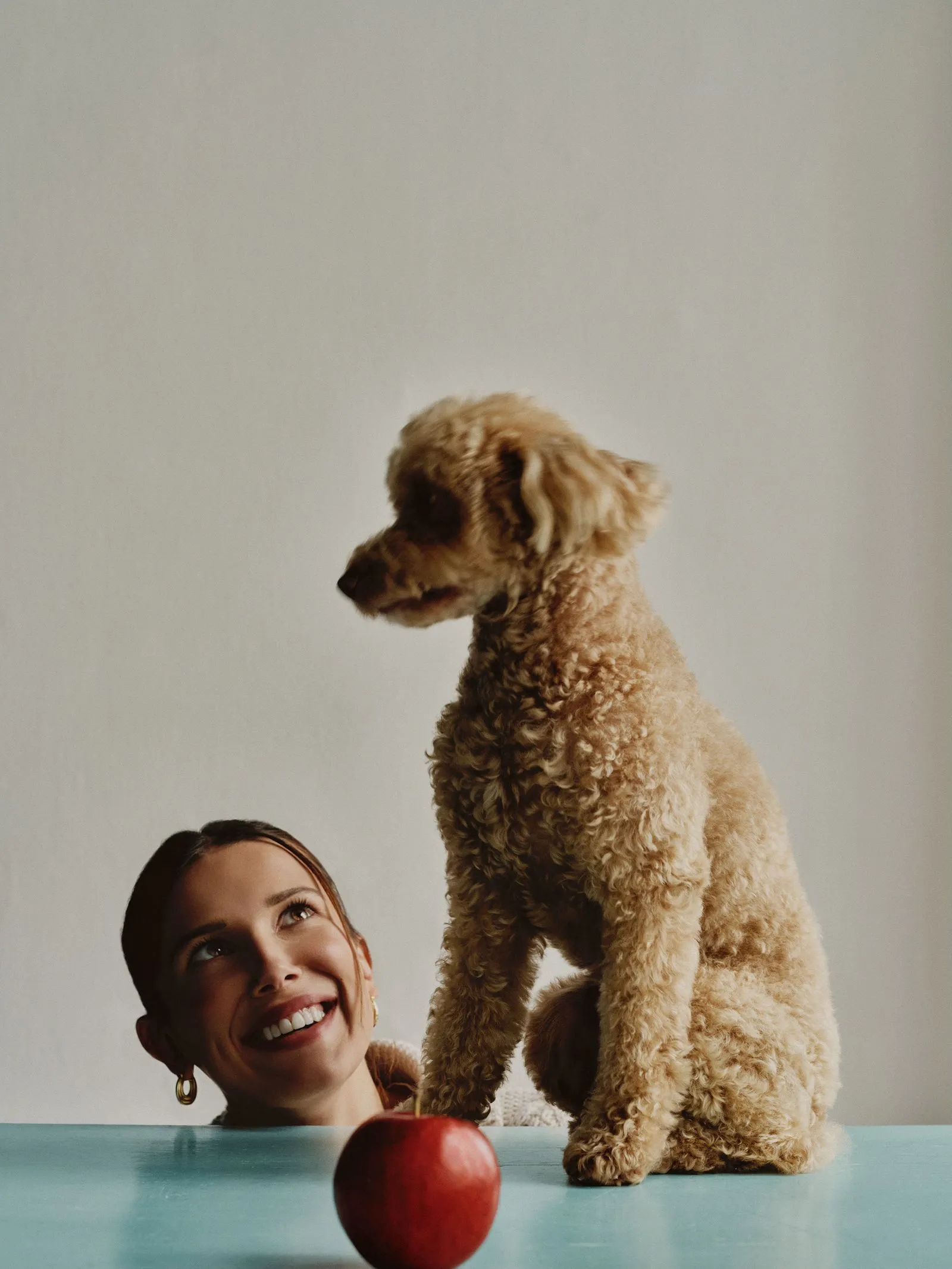 Woman smiling up at a curly brown dog perched on a teal table with a red apple in the foreground.
