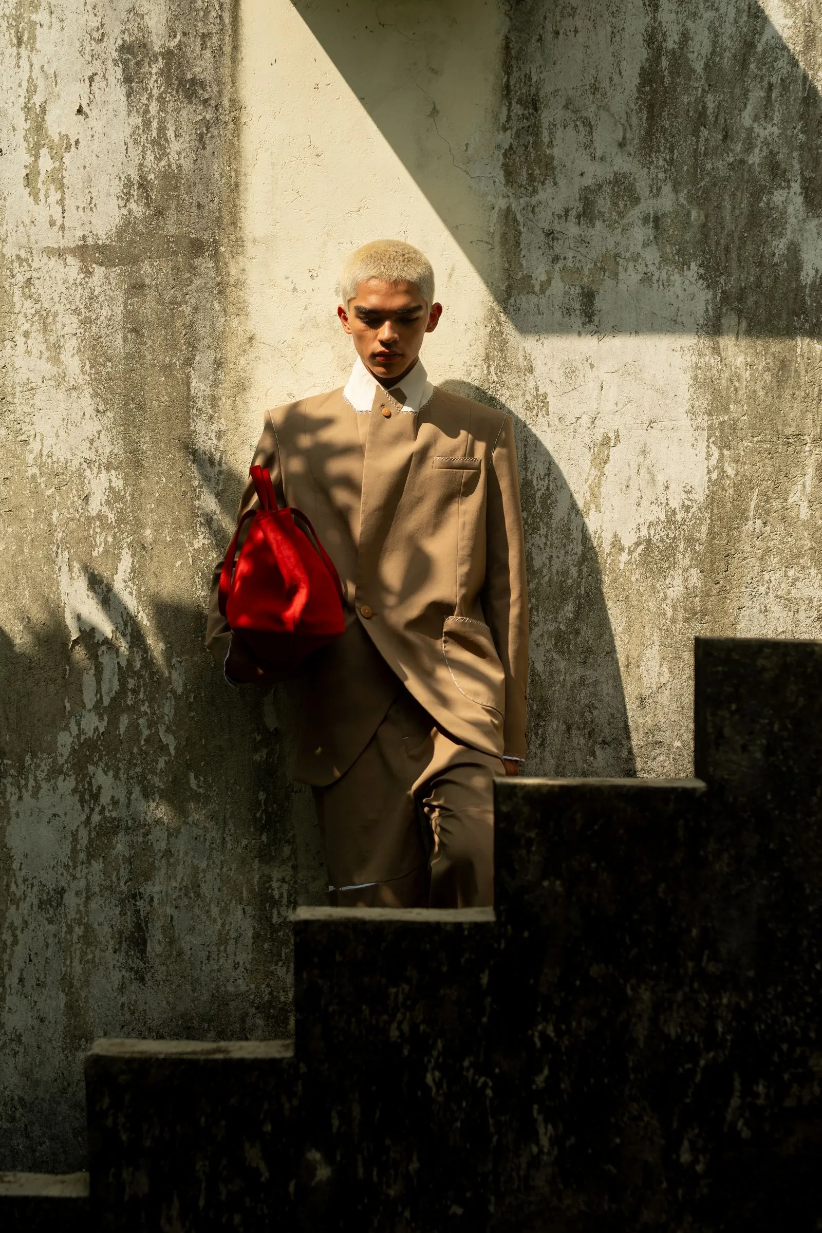 Person with bleached blonde hair in a tan suit holding a red bag, standing beside a weathered wall and stairs in shadows.