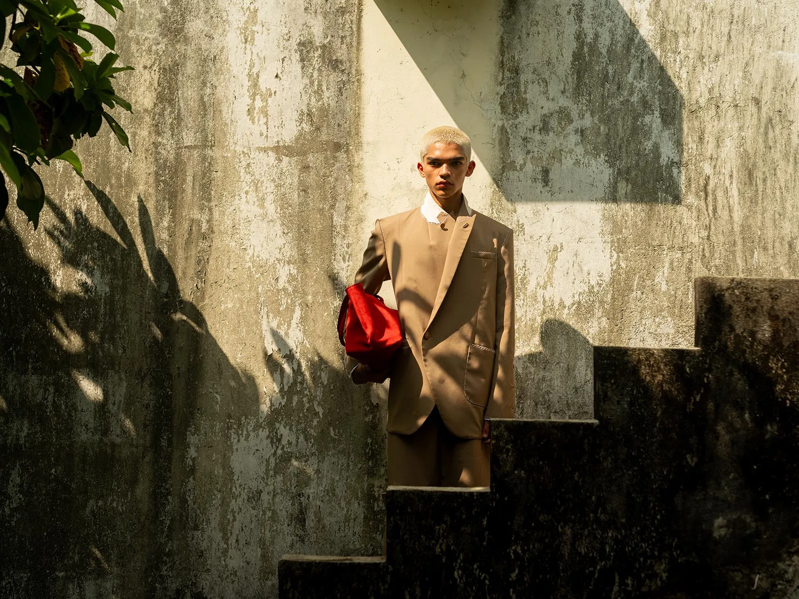 Person with bleach-blond hair wearing a beige suit, holding a red bag, standing on outdoor stairs against a weathered wall.