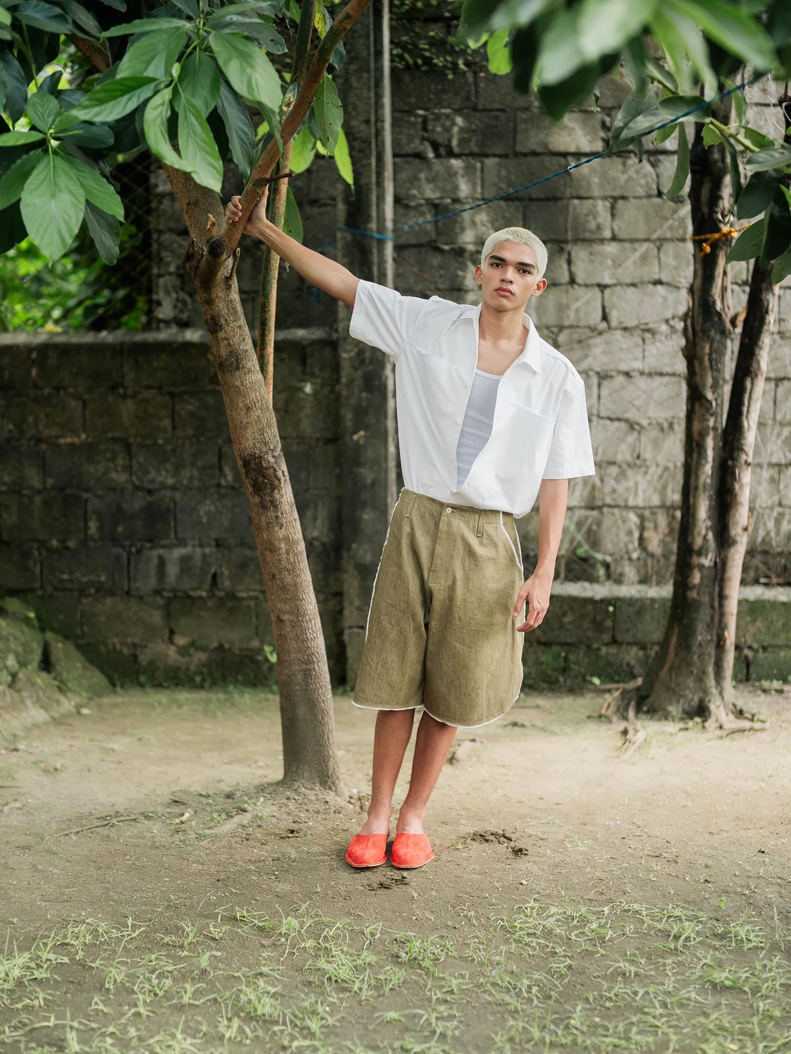 Young person with bleached blond hair stands beside a tree outdoors, wearing a white short-sleeve shirt, tan shorts, and bright orange shoes, looking at the camera.