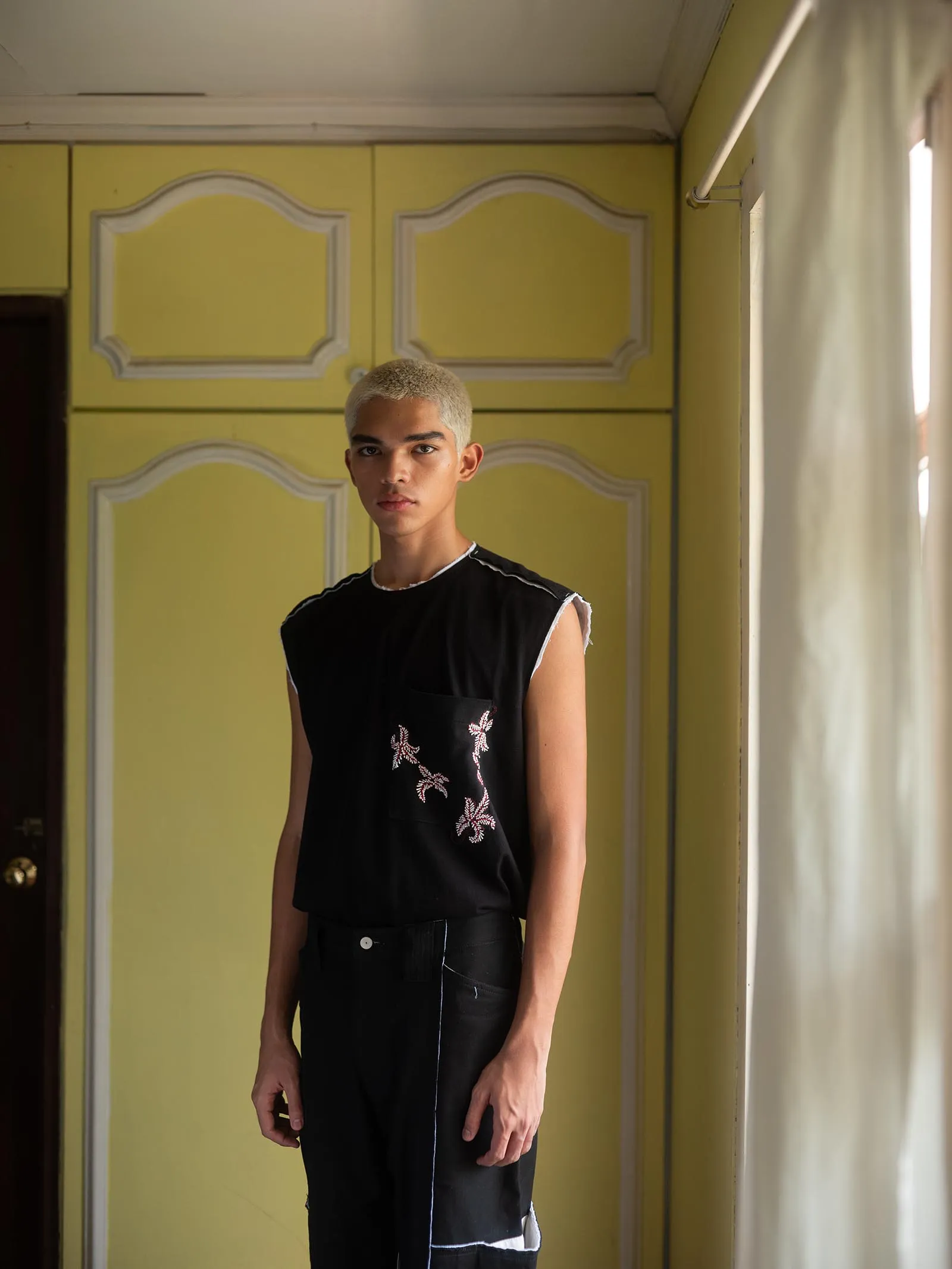 Young man with short blond hair stands indoors in a yellow room, wearing a black sleeveless shirt with white embroidery.