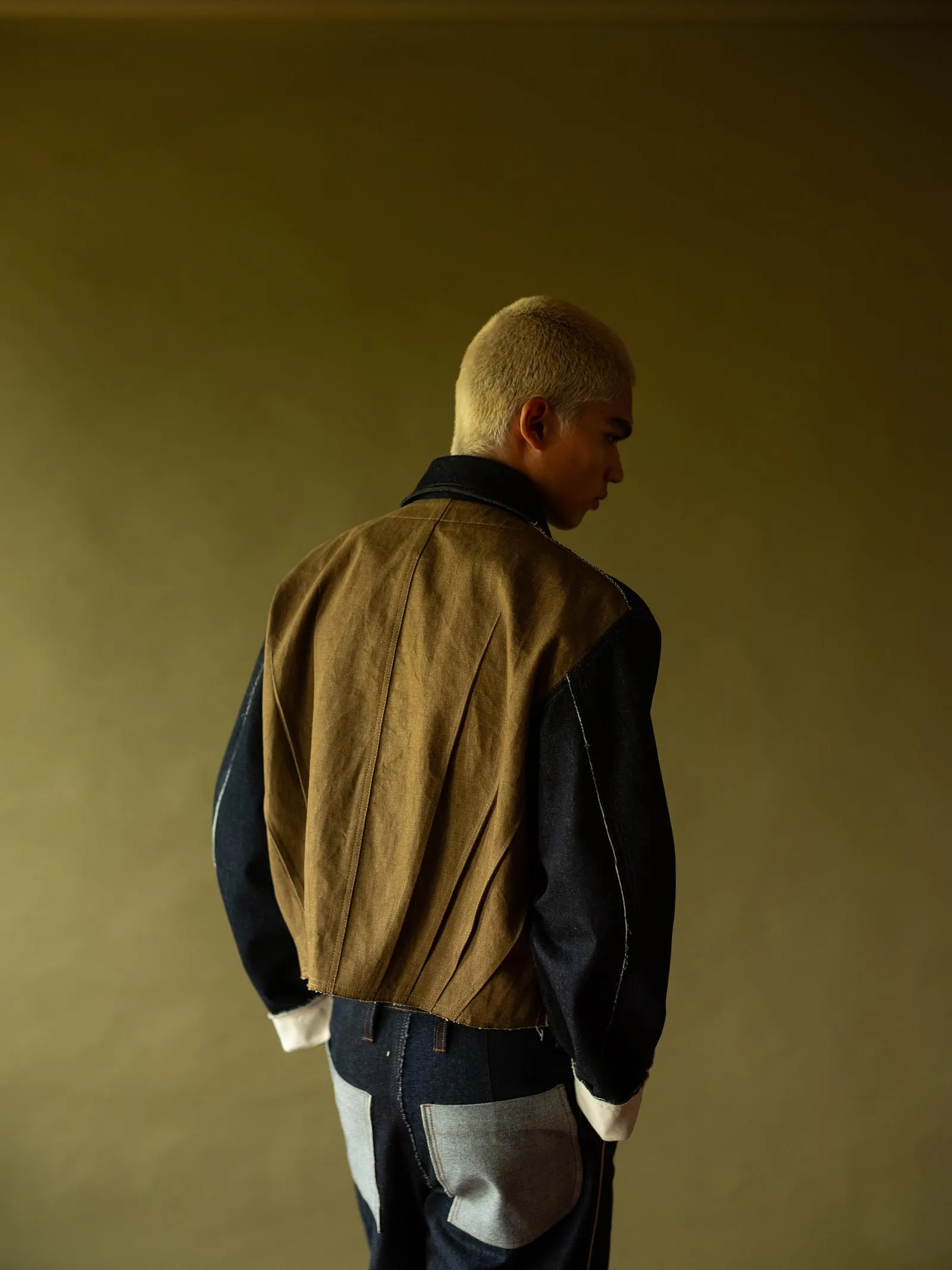 Back view of a person with short blond hair wearing a brown and black color-block jacket, standing against a muted yellow-brown background.