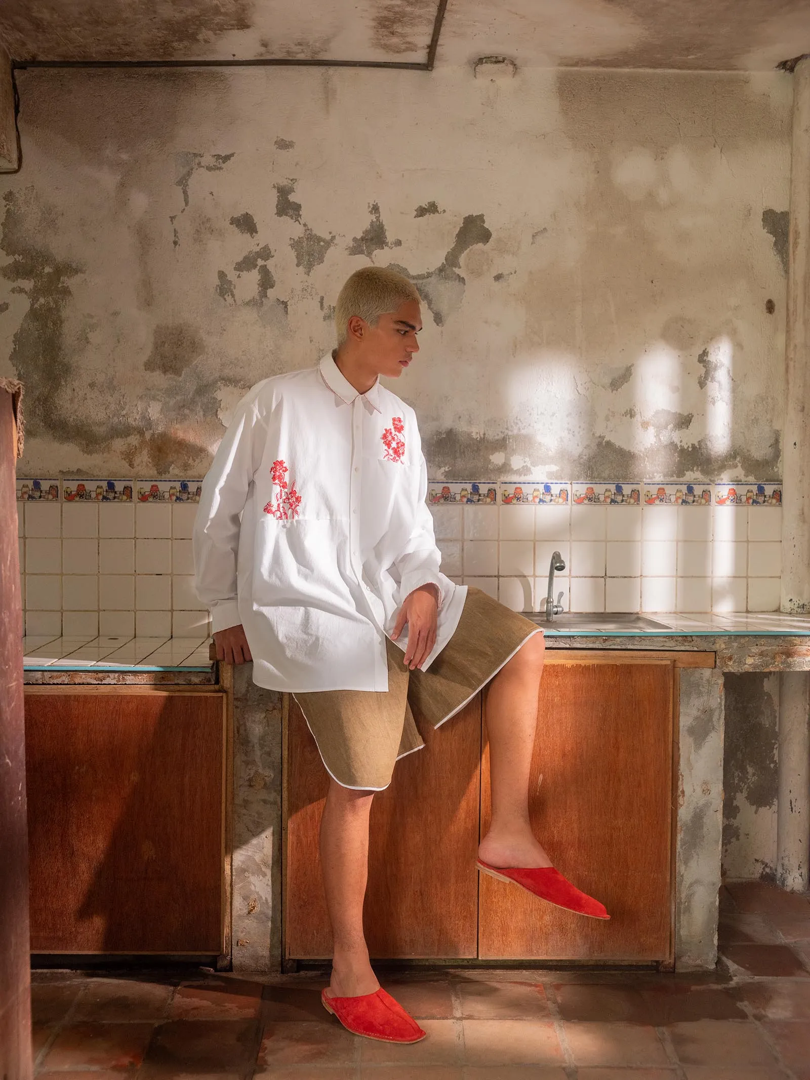 Person with bleached hair wearing a white embroidered shirt and beige wrap skirt sits on a wooden cabinet in a dilapidated kitchen with peeling walls and tiles.
