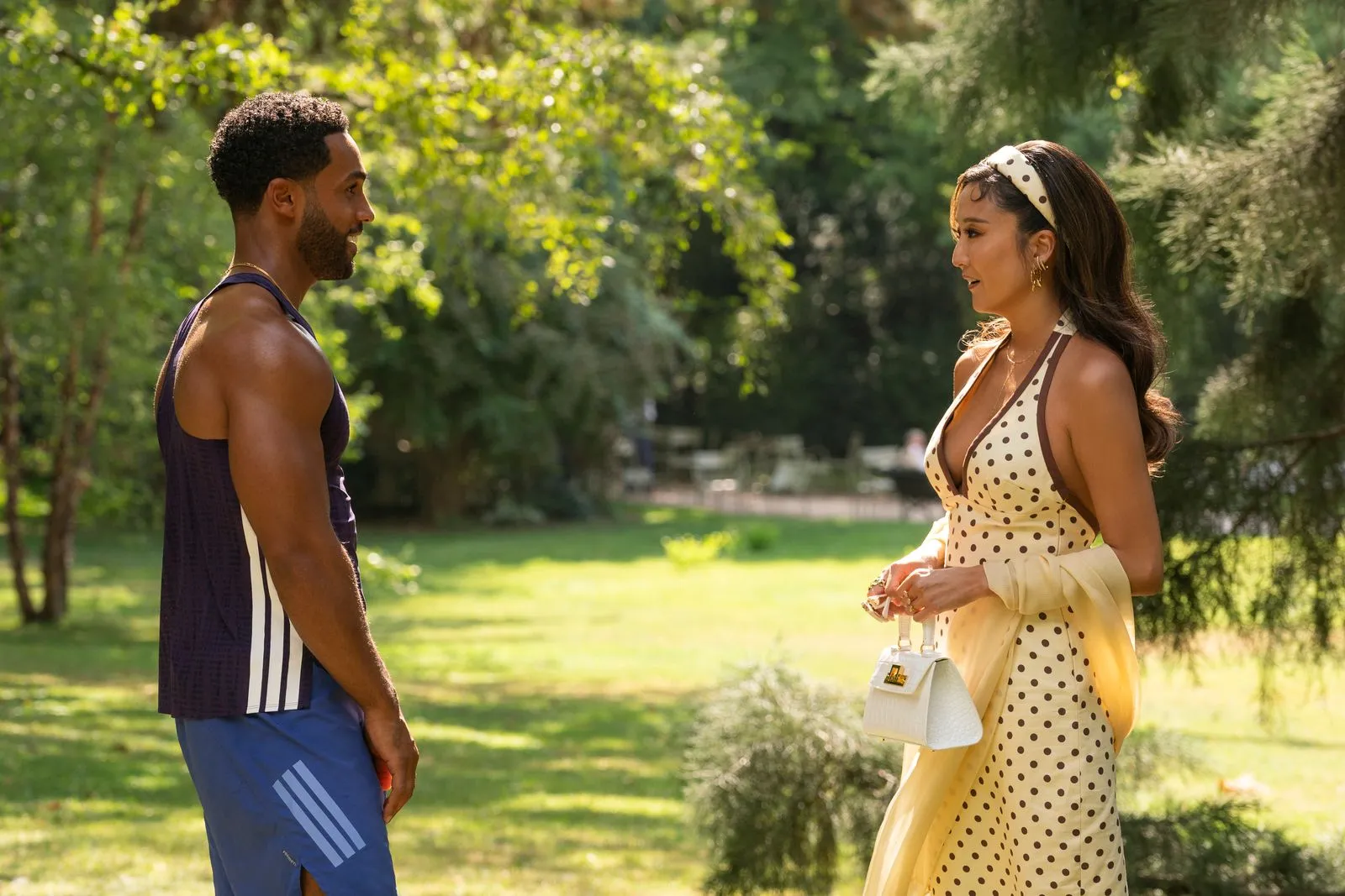 Two people stand in a sunlit park, facing each other as they chat (man in athletic wear, woman in a cream polka-dot dress with a white purse).