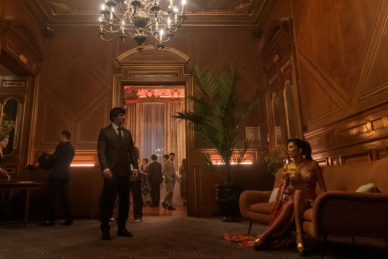 Man in a dark suit stands in a dimly lit, ornate room with a chandelier; a woman in a gold gown sits on an orange sofa to the right.
