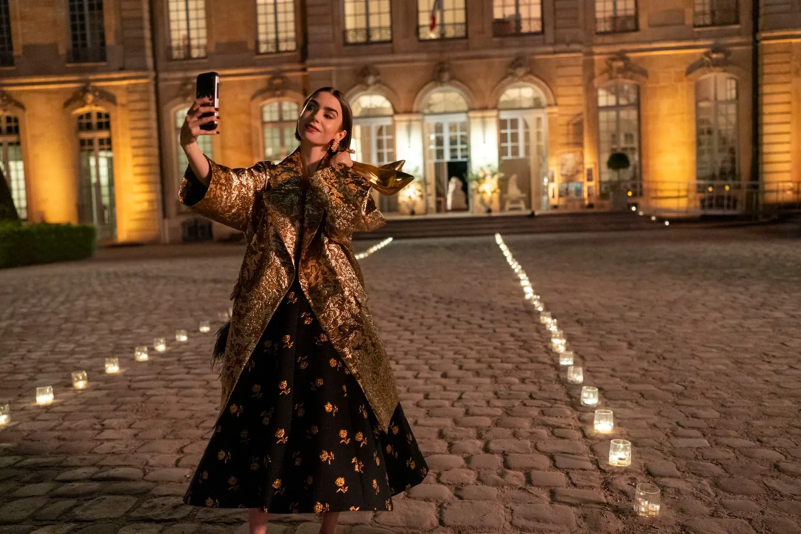Woman in a gold brocade coat taking a selfie in a candlelit courtyard at night.