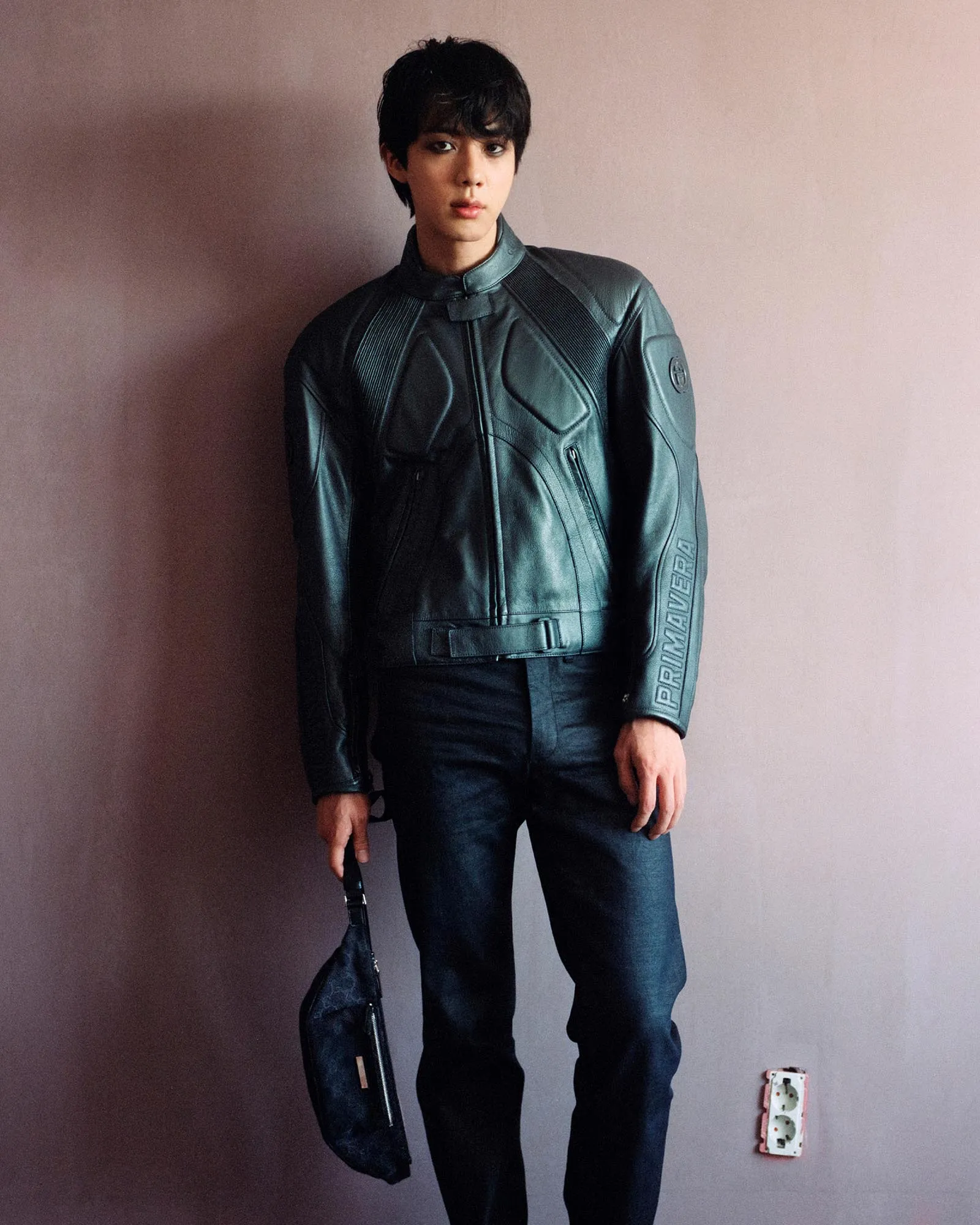 Young man in a dark leather motorcycle jacket stands against a pink wall, holding a black strap or bag in one hand.