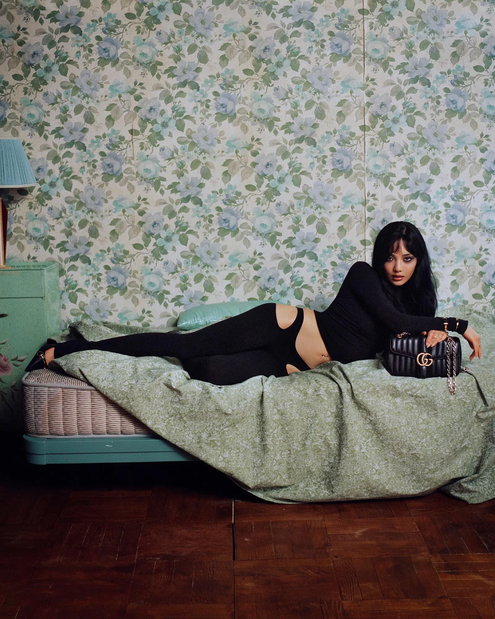Woman reclining on a bed in a room with blue floral wallpaper, wearing black clothing and holding a black purse with a chain strap.
