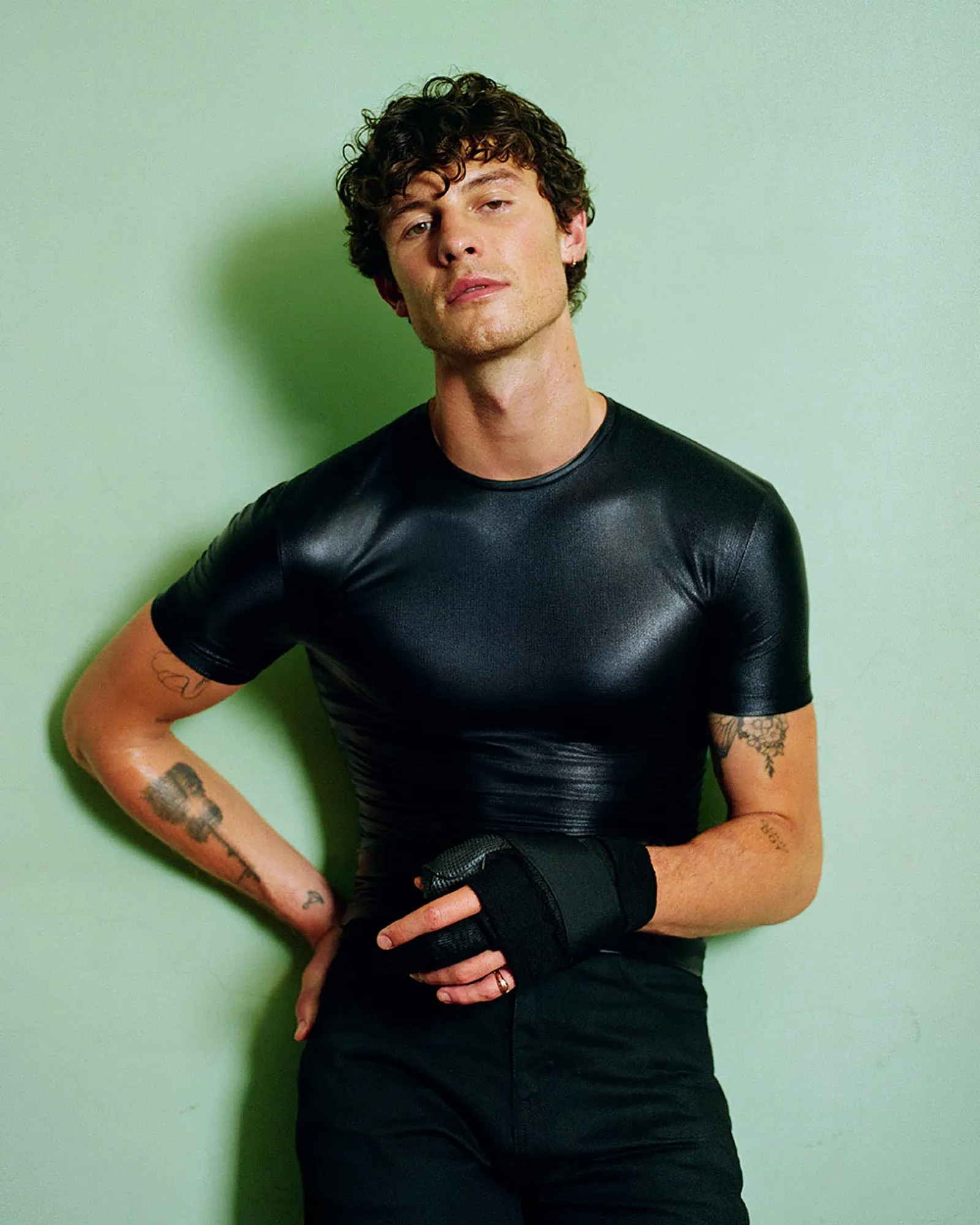 Man in a black, tight athletic shirt leaning against a light green wall, arms crossed.