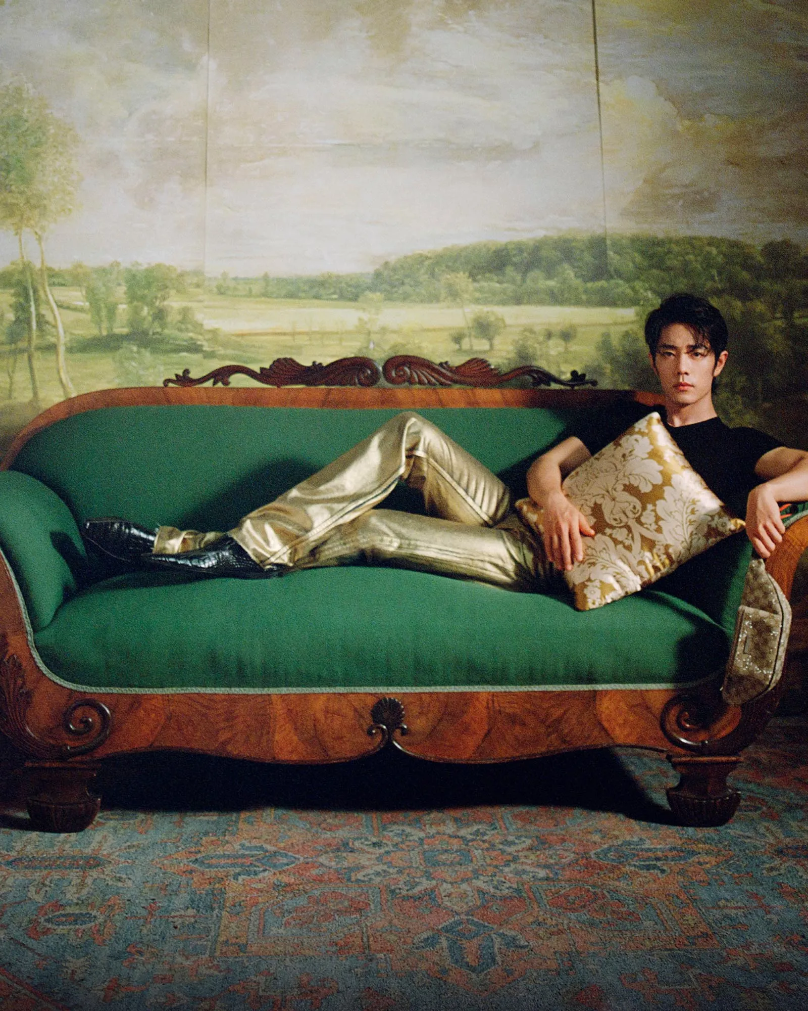 Young man reclining on a vintage green sofa, holding a floral pillow, with a landscape mural in the background.