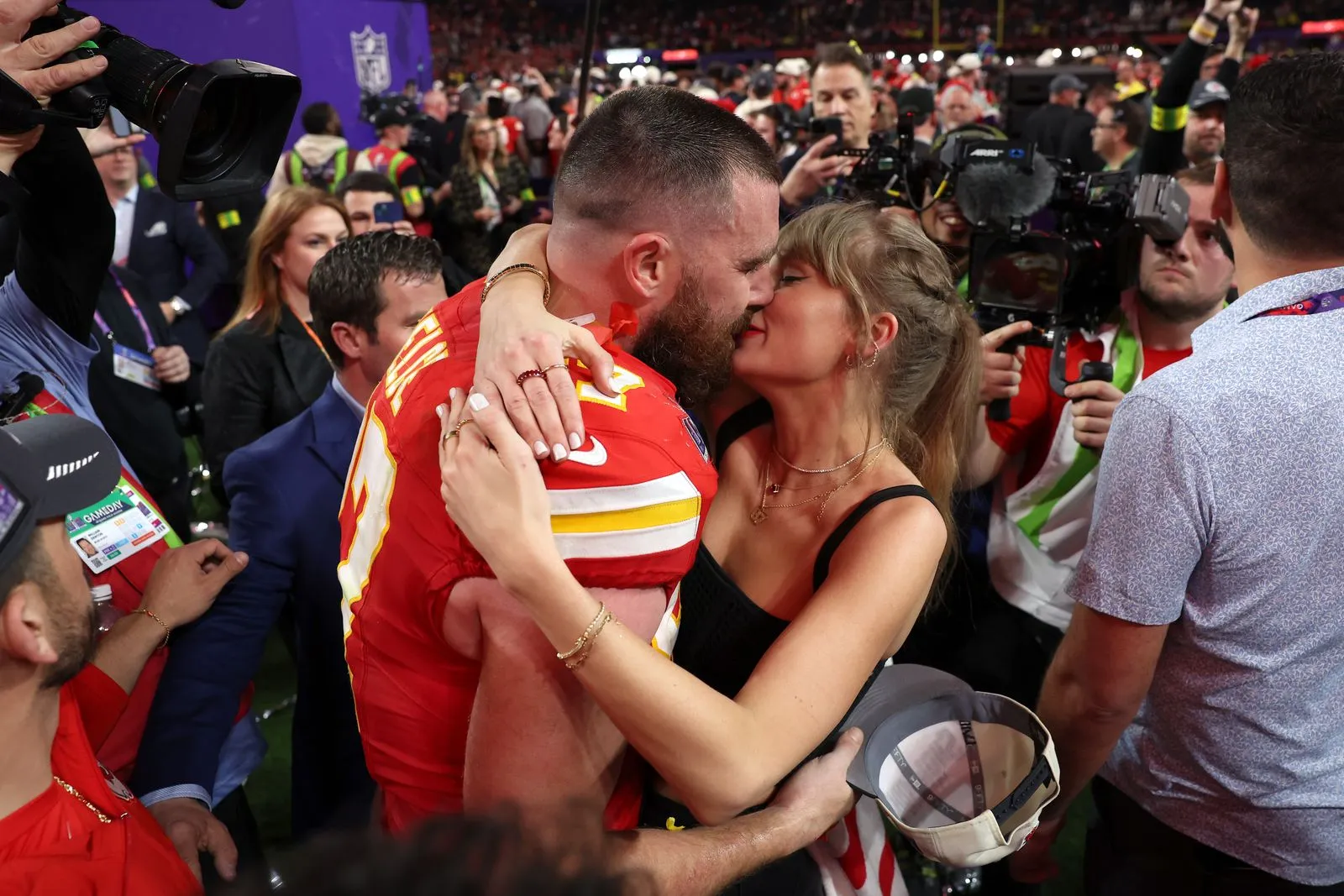 Athlete in red jersey kissing a woman amid a crowded media scrum with cameras and reporters nearby at a stadium event.