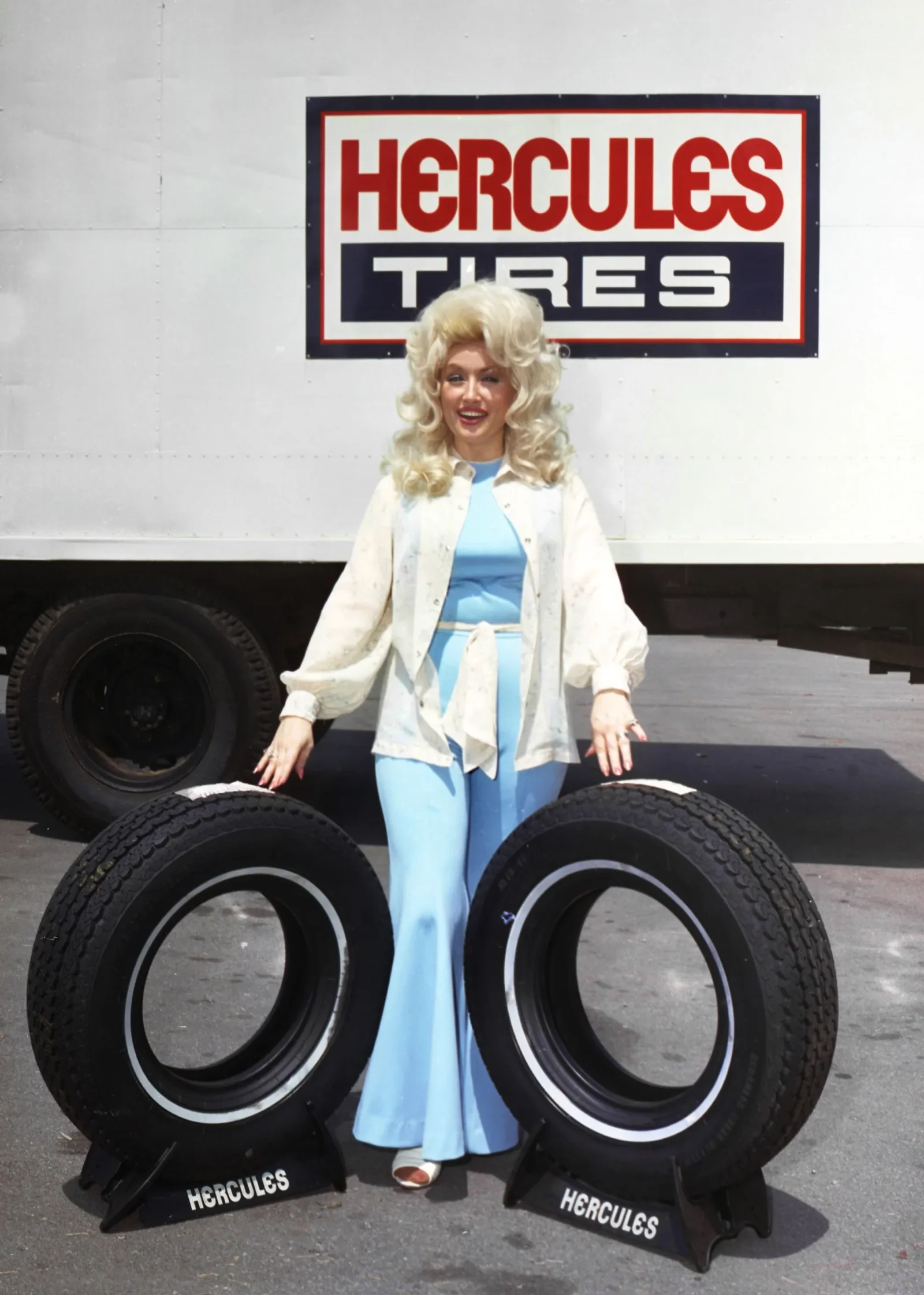 Woman with big blonde hair in a light blue outfit stands among large tires in front of a Hercules Tires sign.