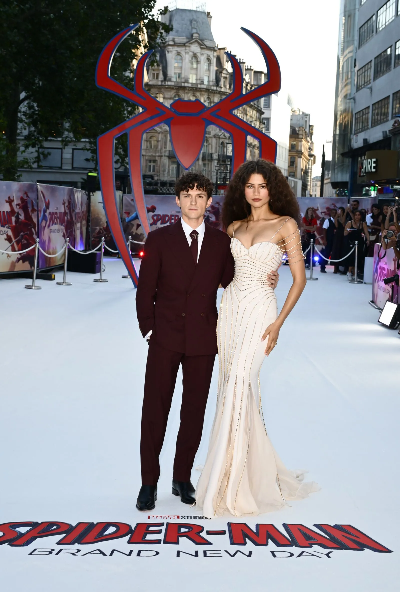 Tom Holland and Zendaya pose on a white carpet at a Spider-Man premiere with a large red-and-blue Spider-Man emblem behind them.