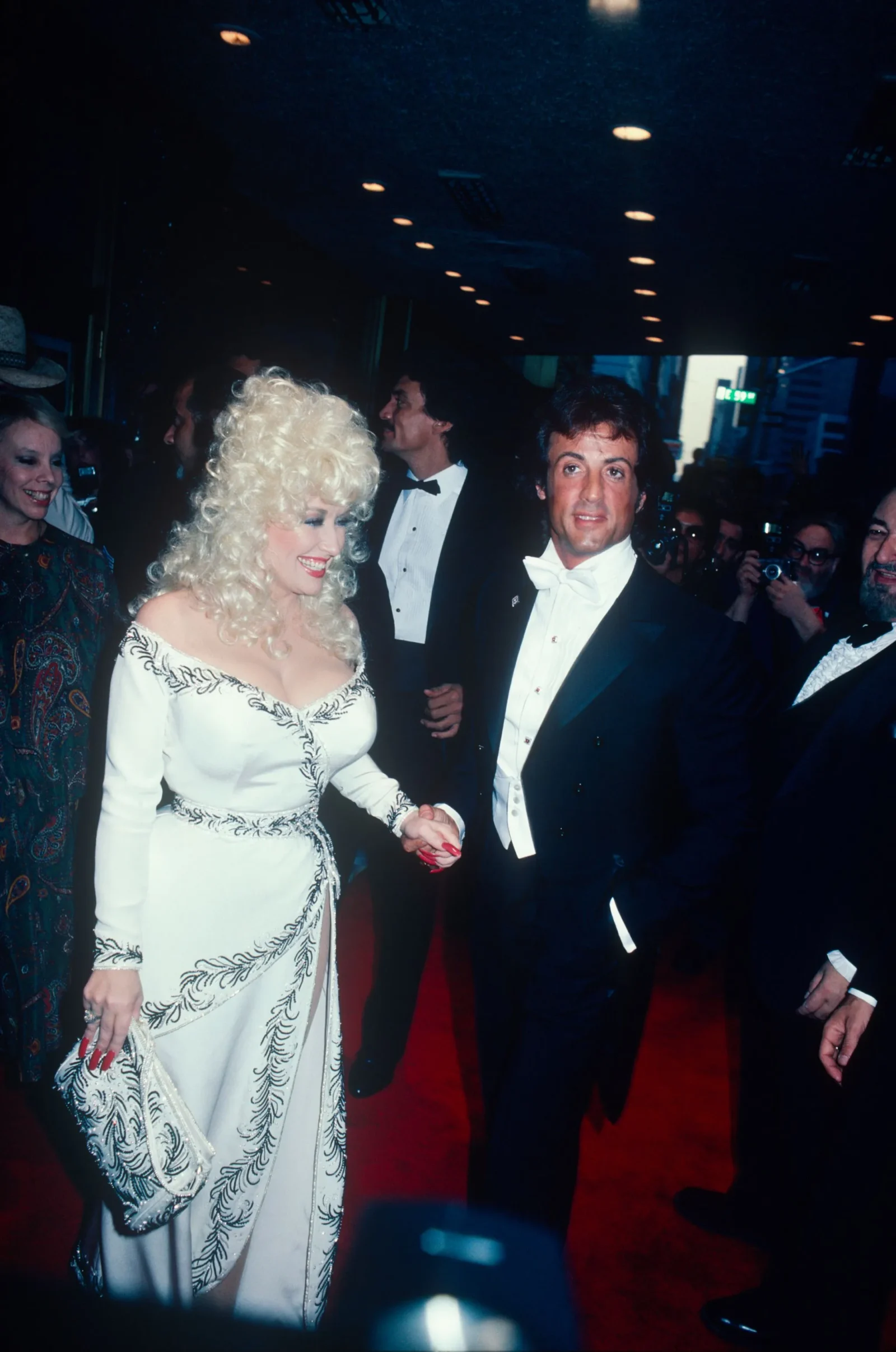 Celebrity couple on a red carpet; woman in a white gown with platinum curls and clutch, man in a black tuxedo beside her.