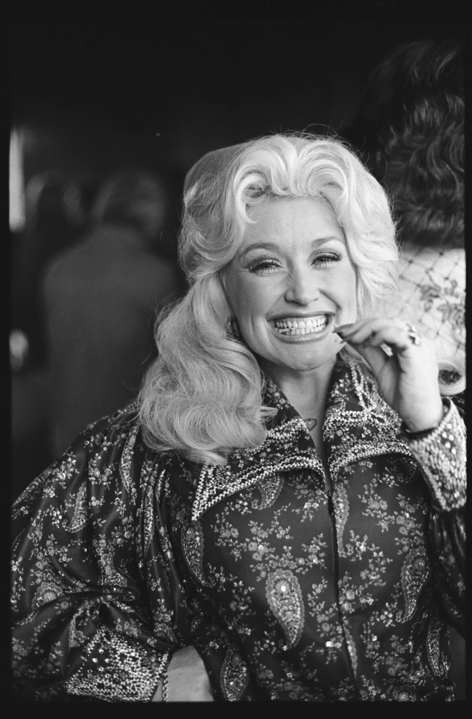 Black-and-white portrait of a smiling blonde woman wearing a patterned, ornate jacket or blouse, looking toward the camera.