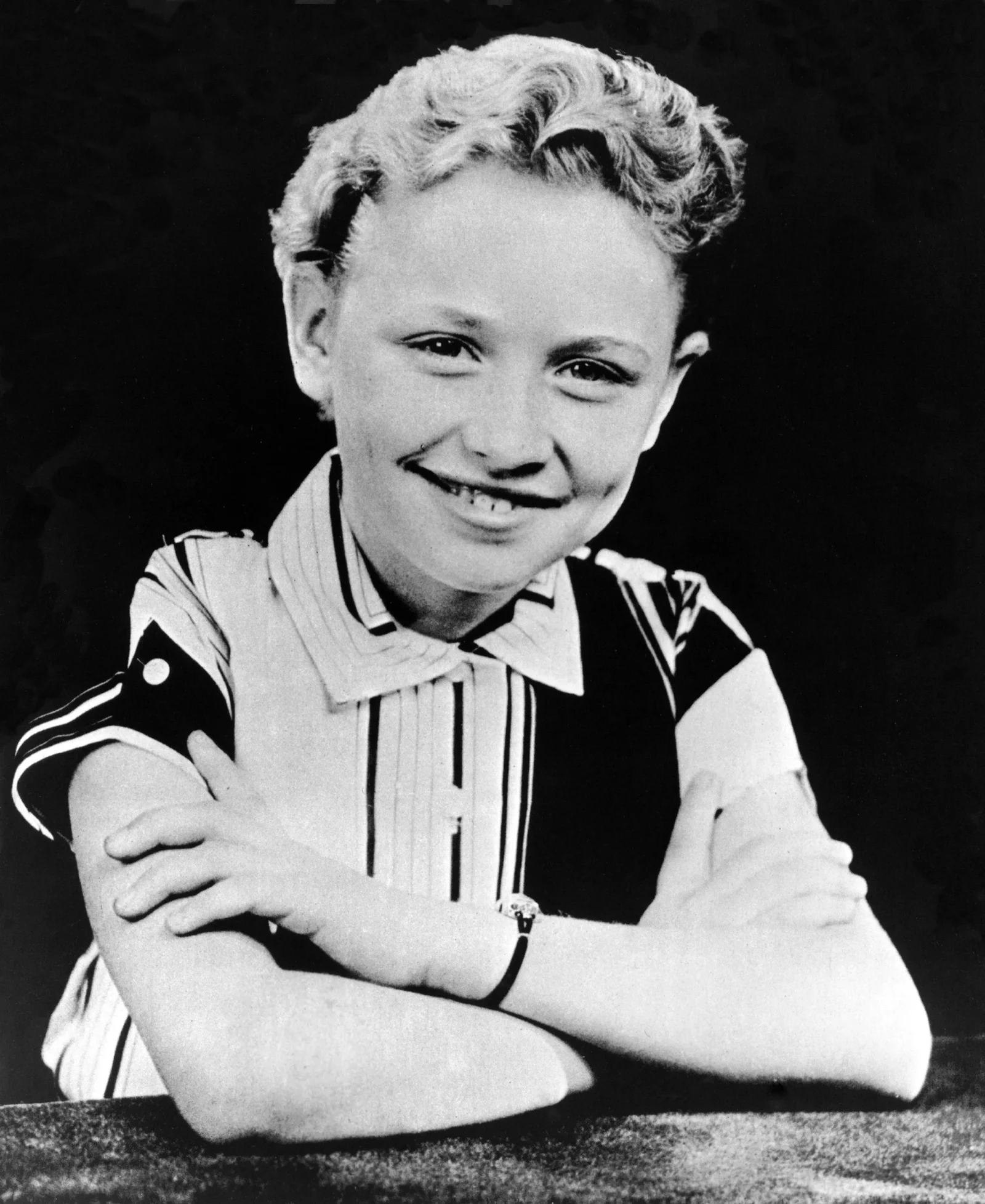 Smiling boy with short, wavy hair wearing a striped shirt, arms crossed on a table against a dark background (black-and-white photo).