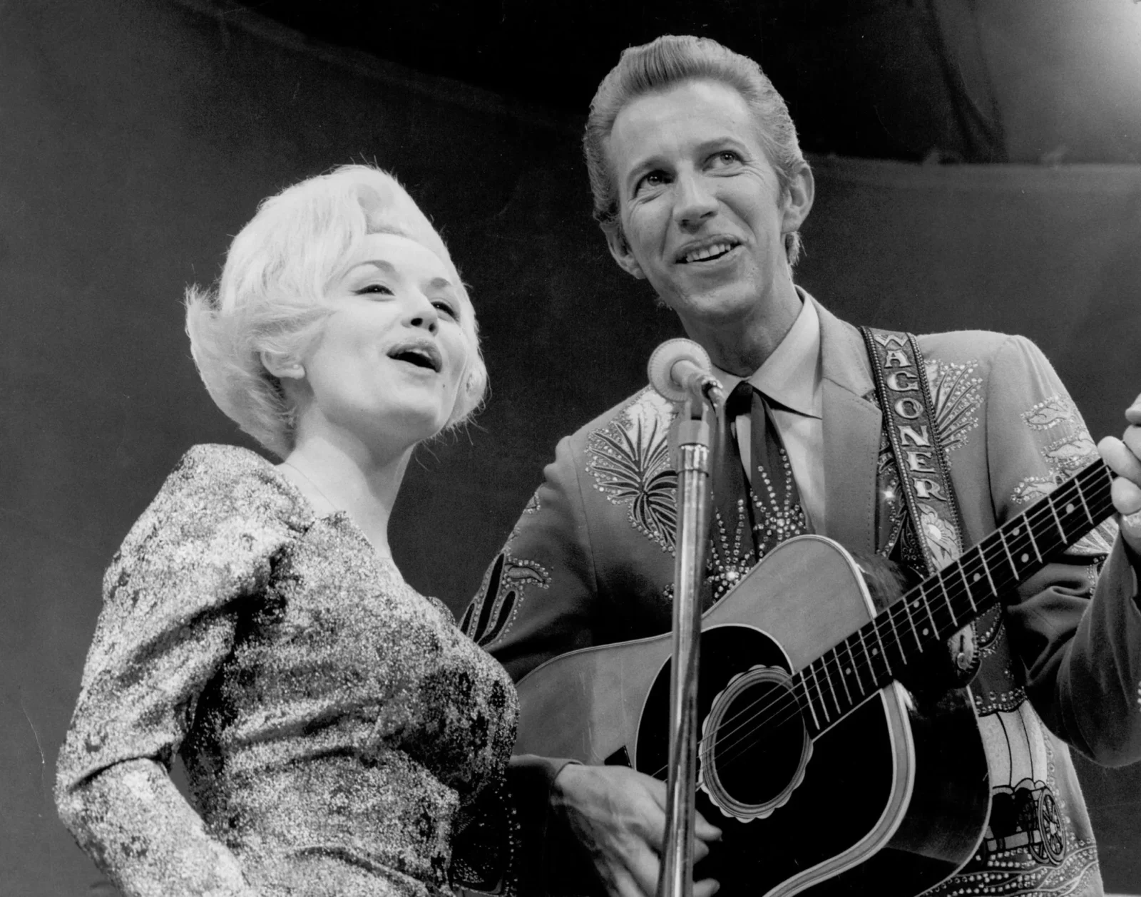 Black-and-white photo of Dolly Parton singing beside Porter Wagoner, who plays an acoustic guitar on stage.
