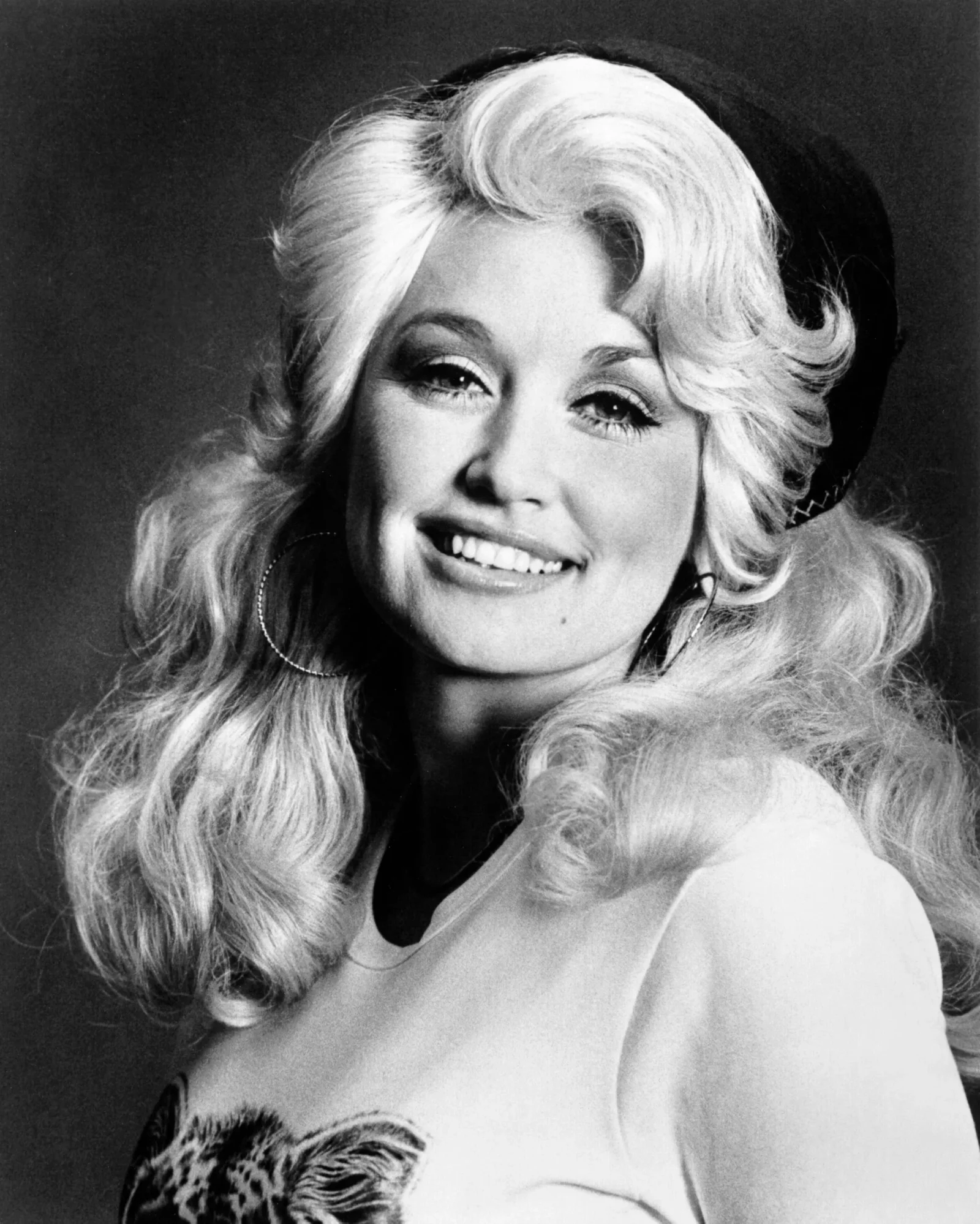 Black-and-white close-up portrait of a smiling blonde woman with hoop earrings and voluminous curls in a studio setting.