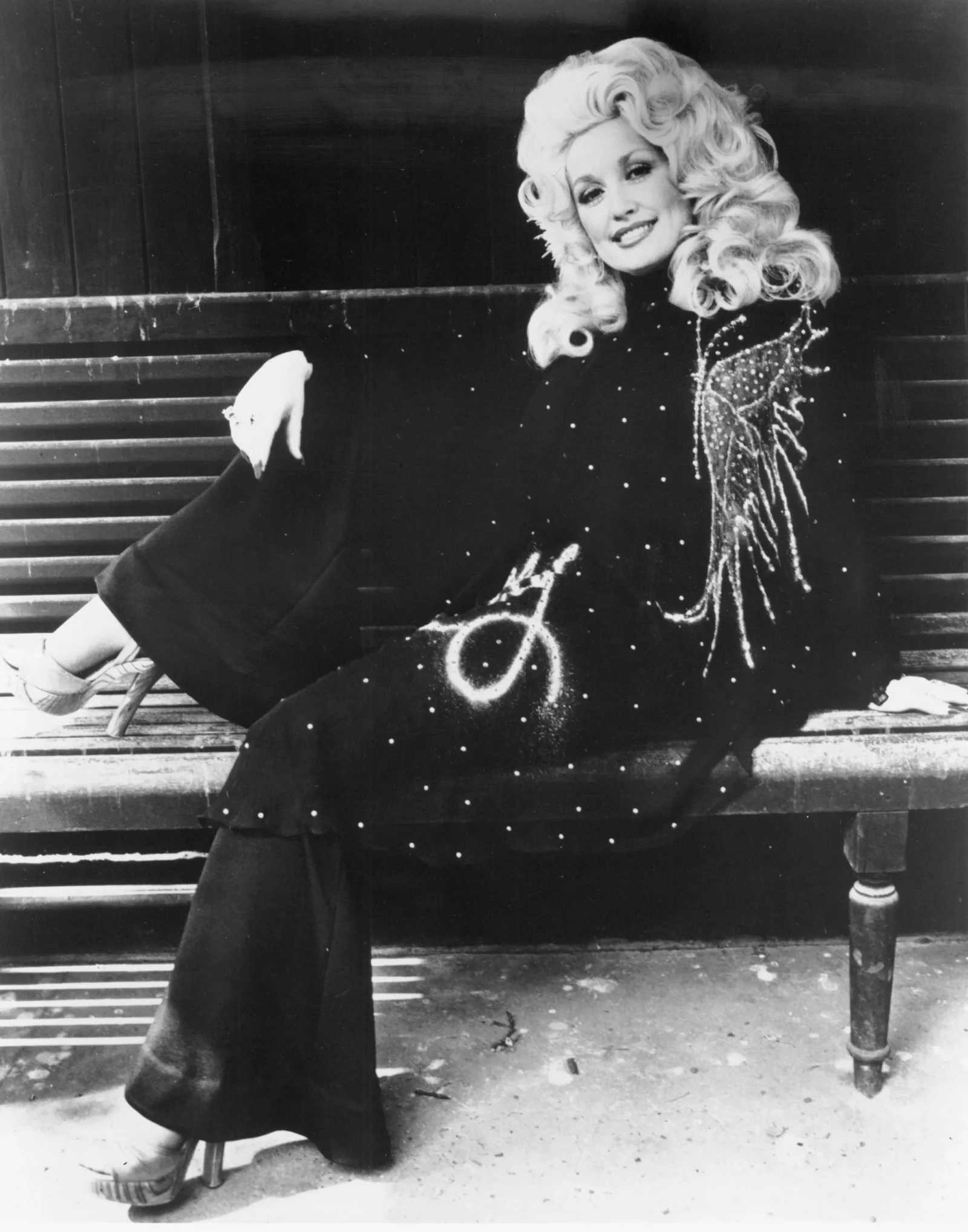 Black-and-white photo of a smiling woman with voluminous blonde curls, wearing a sparkly black outfit, seated on a wooden bench outside.