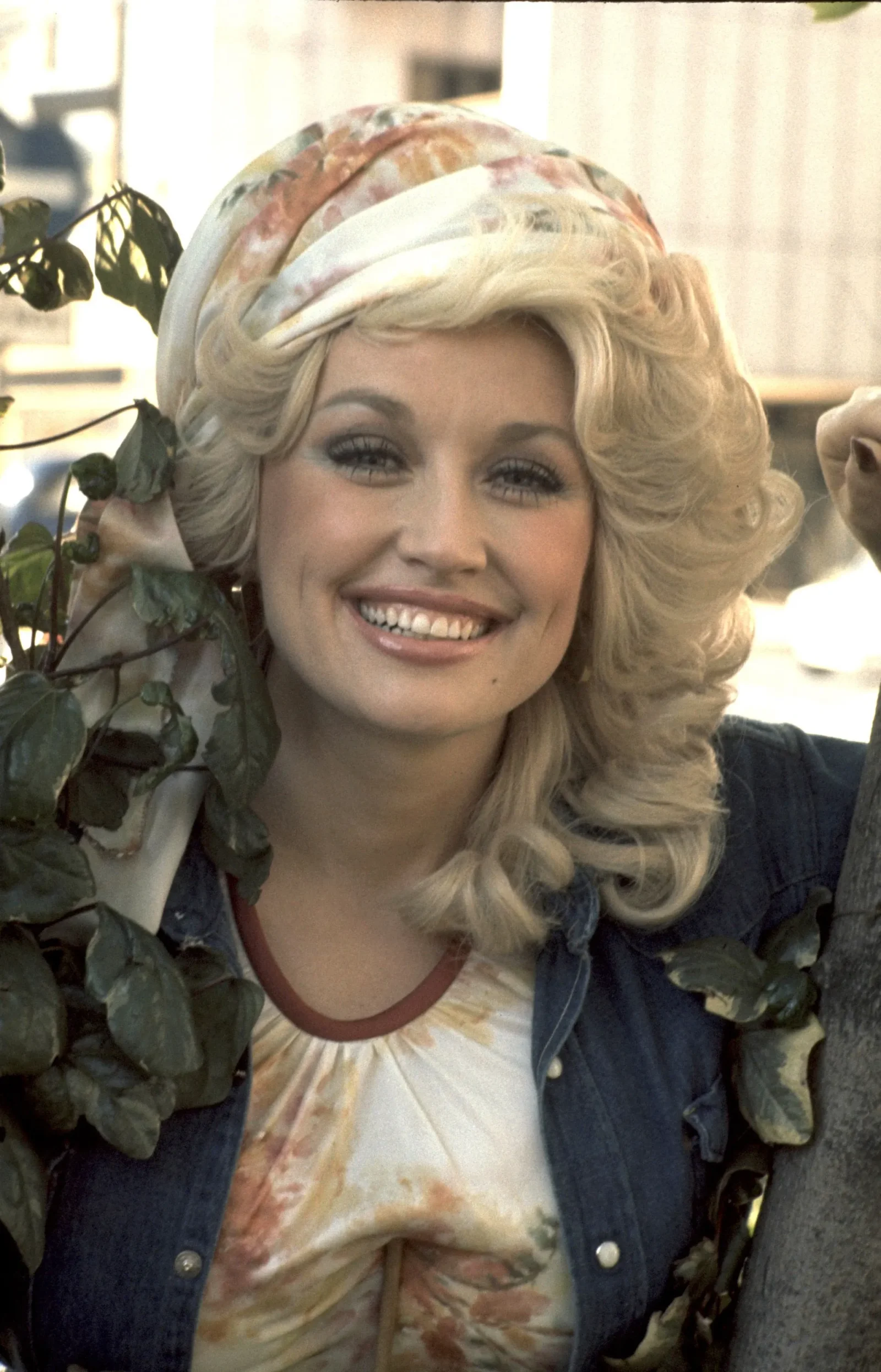 Smiling blonde woman in a floral headscarf and denim jacket, surrounded by ivy leaves outdoors.