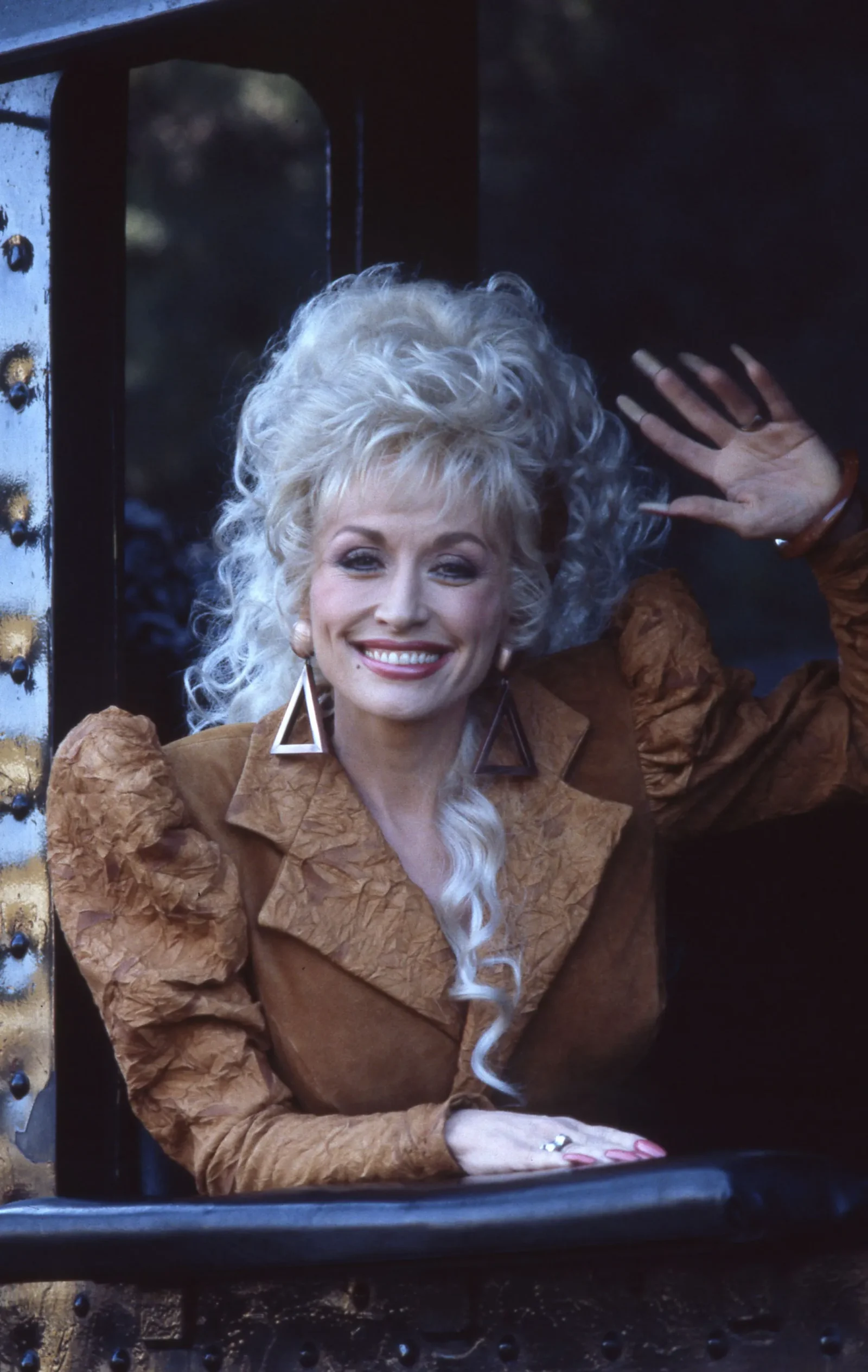 Smiling woman with platinum blonde hair, wearing a brown textured coat, waving from a dark train window frame.