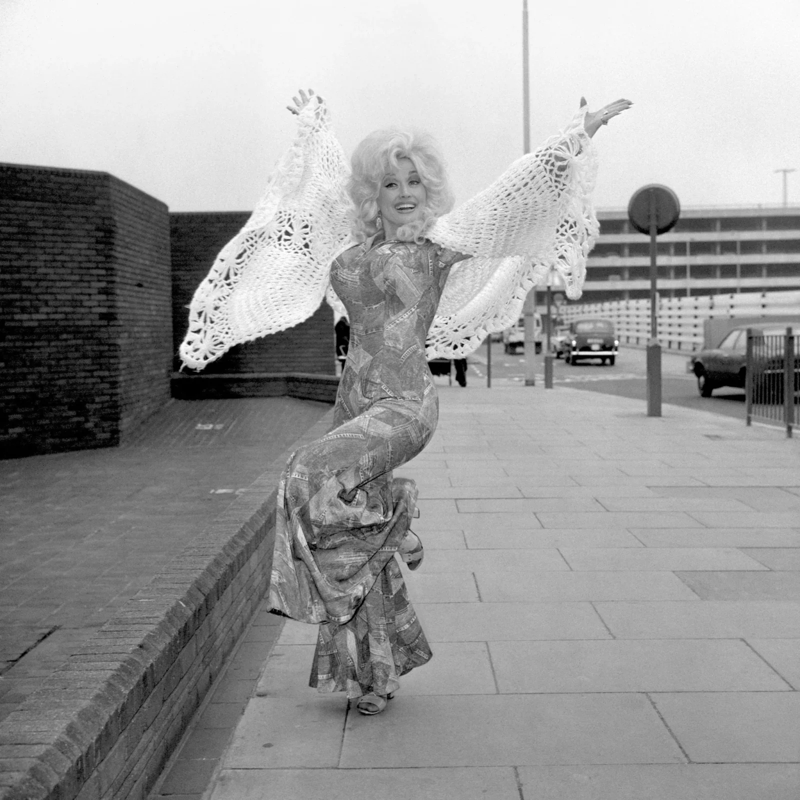 Woman in patterned dress wearing a large crocheted shawl, arms raised in a joyful pose outdoors in an urban setting.