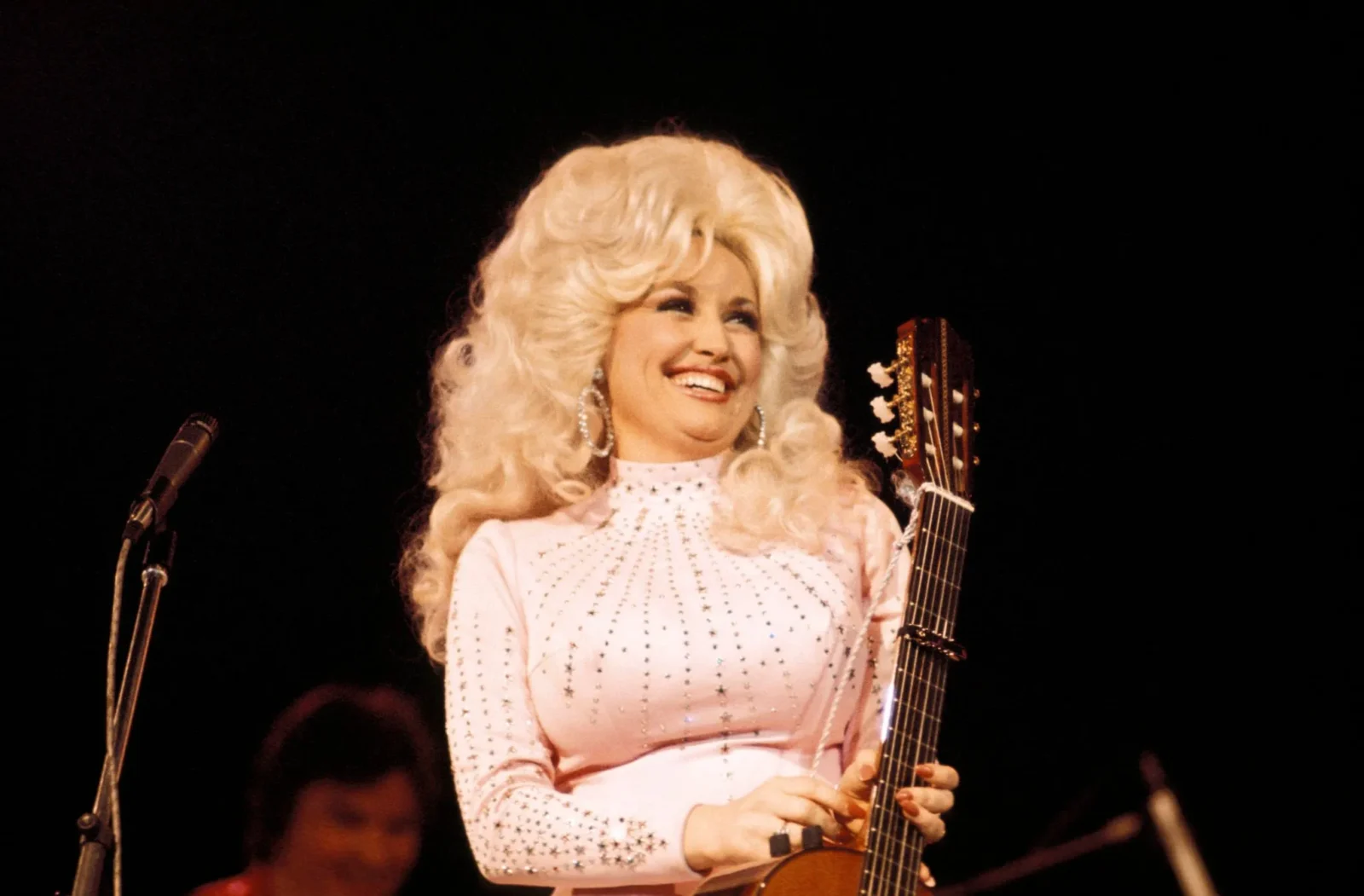 Female singer with long platinum blonde hair in a pale pink studded dress, holding a guitar on stage with a smile.