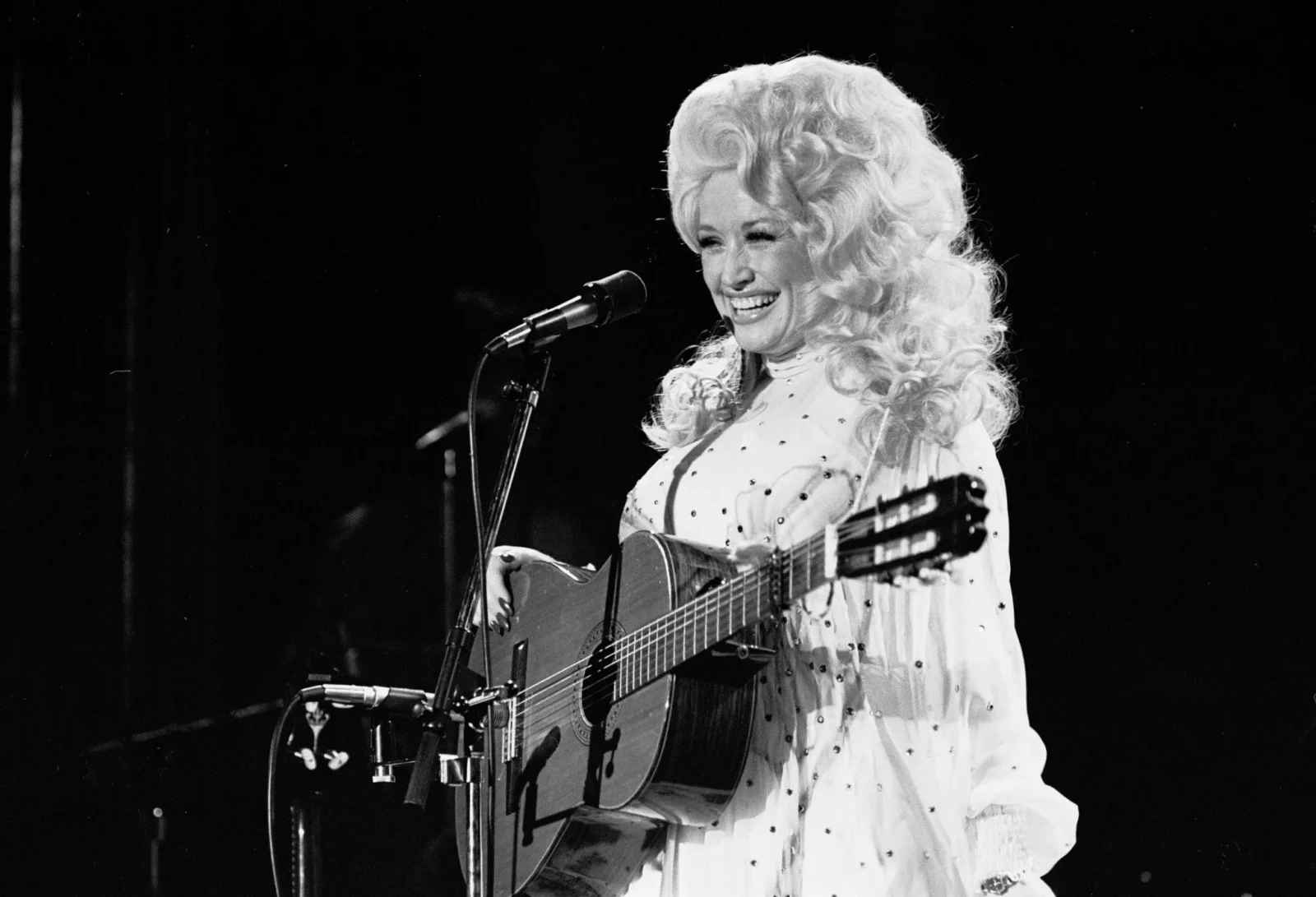 Female singer with long curly blonde hair, performing on stage with an acoustic guitar and microphone.