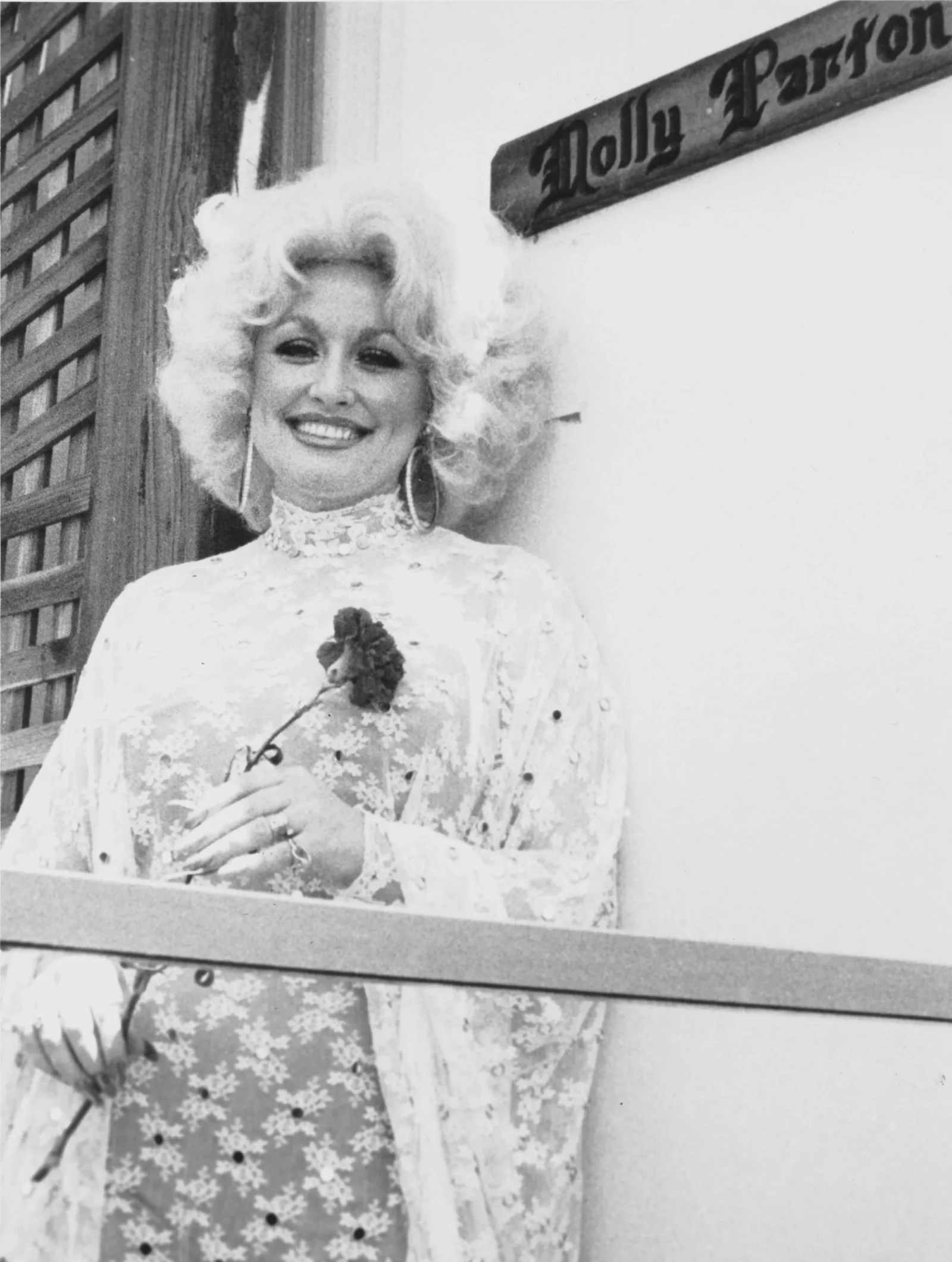 Smiling woman with blonde curly hair wearing a floral lace dress, standing near a wooden lattice outdoors.