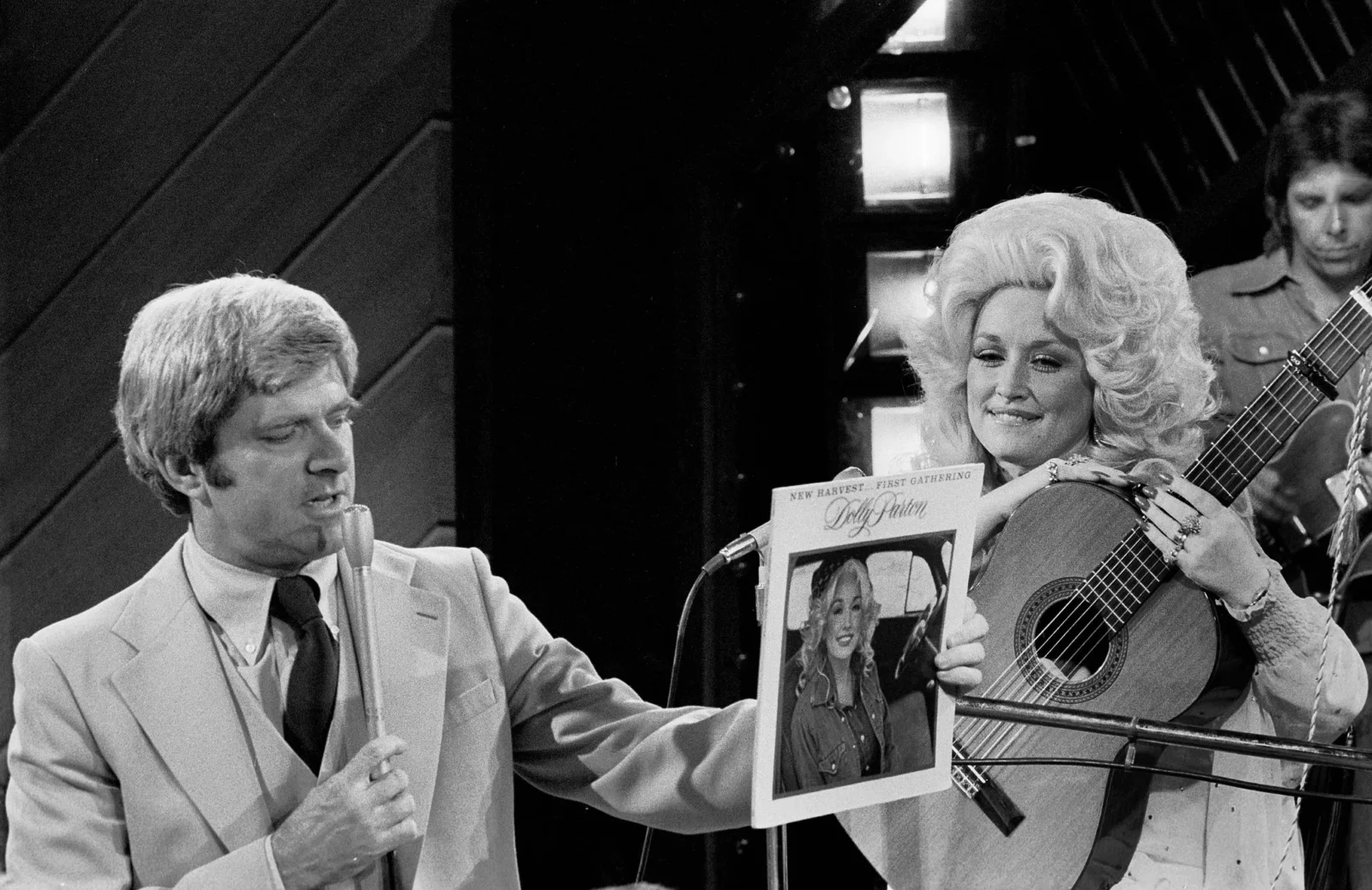 Male singer in a light suit sings into a microphone while a woman with big, styled hair plays a guitar and reads a magazine on stage (black-and-white).