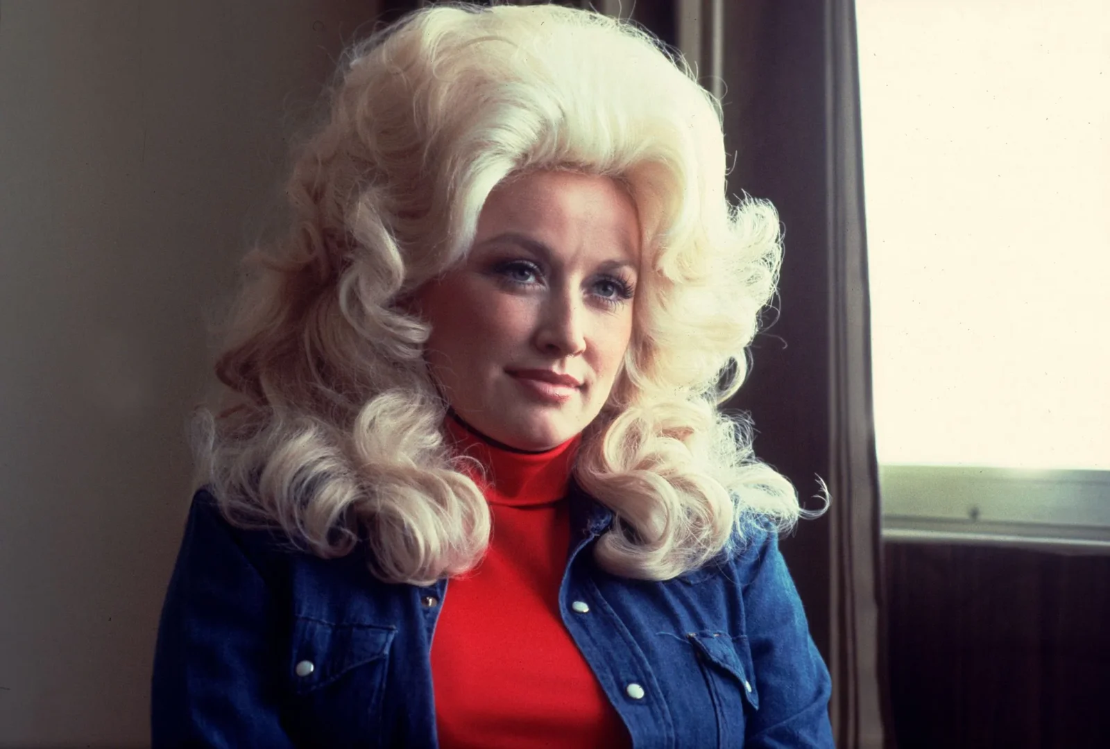 Smiling woman with voluminous platinum blonde hair wearing a red top and blue denim jacket by a window.