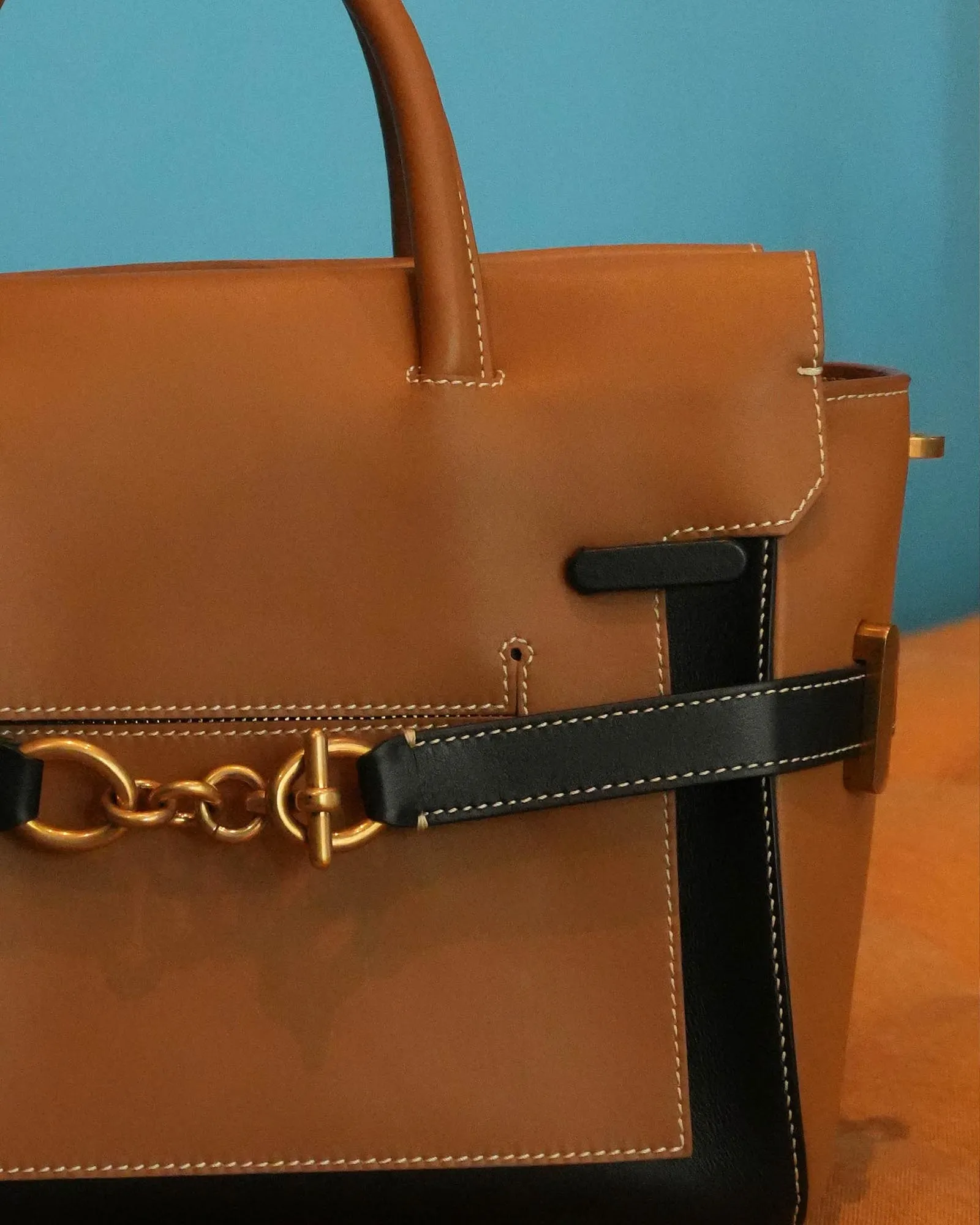 Close-up of a tan leather handbag showing white stitching, a curved handle, and gold hardware against a teal background.