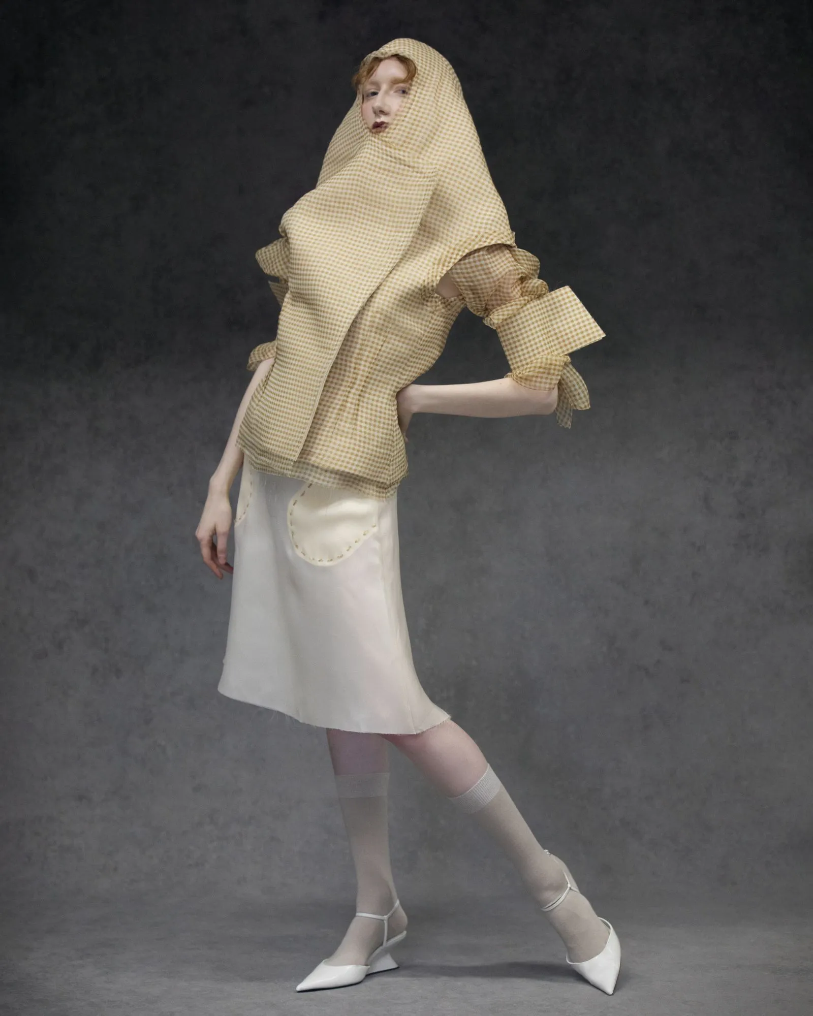 Model in a beige gingham hooded cape with oversized sleeves, white skirt, and white heels against a dark backdrop.