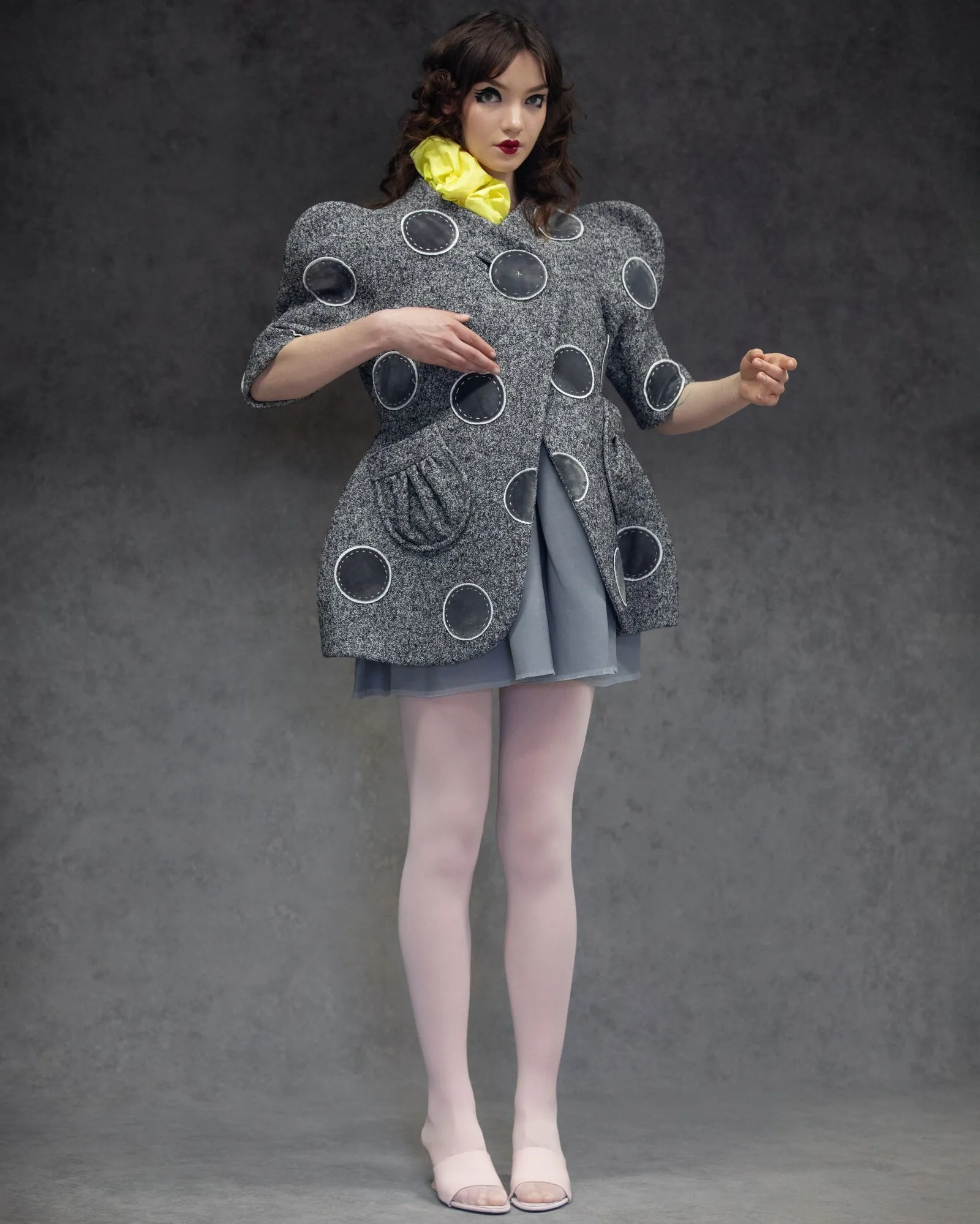 Model wearing a textured gray coat with large circular patches, yellow scarf, and a blue skirt against a dark backdrop.