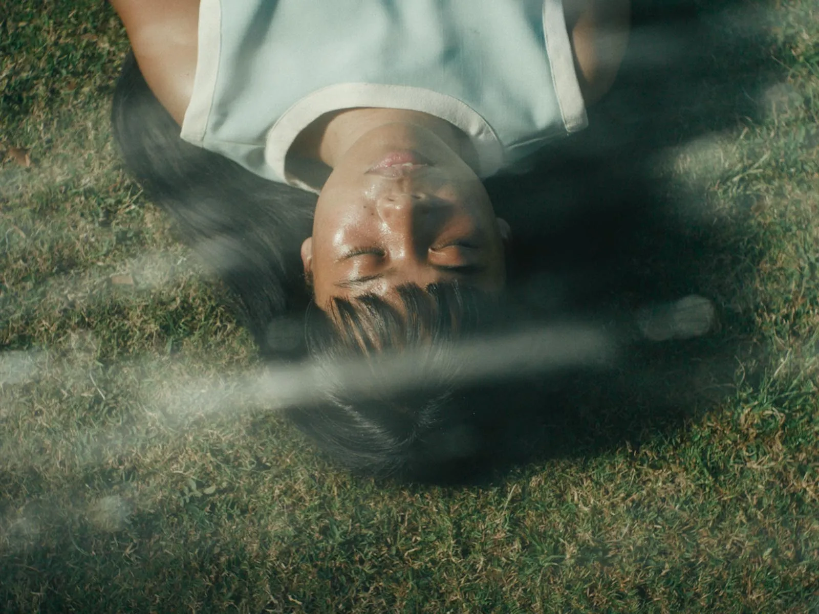 Person lying on grass with eyes closed, viewed from above in a relaxed outdoor scene, with a soft, blurred foreground.