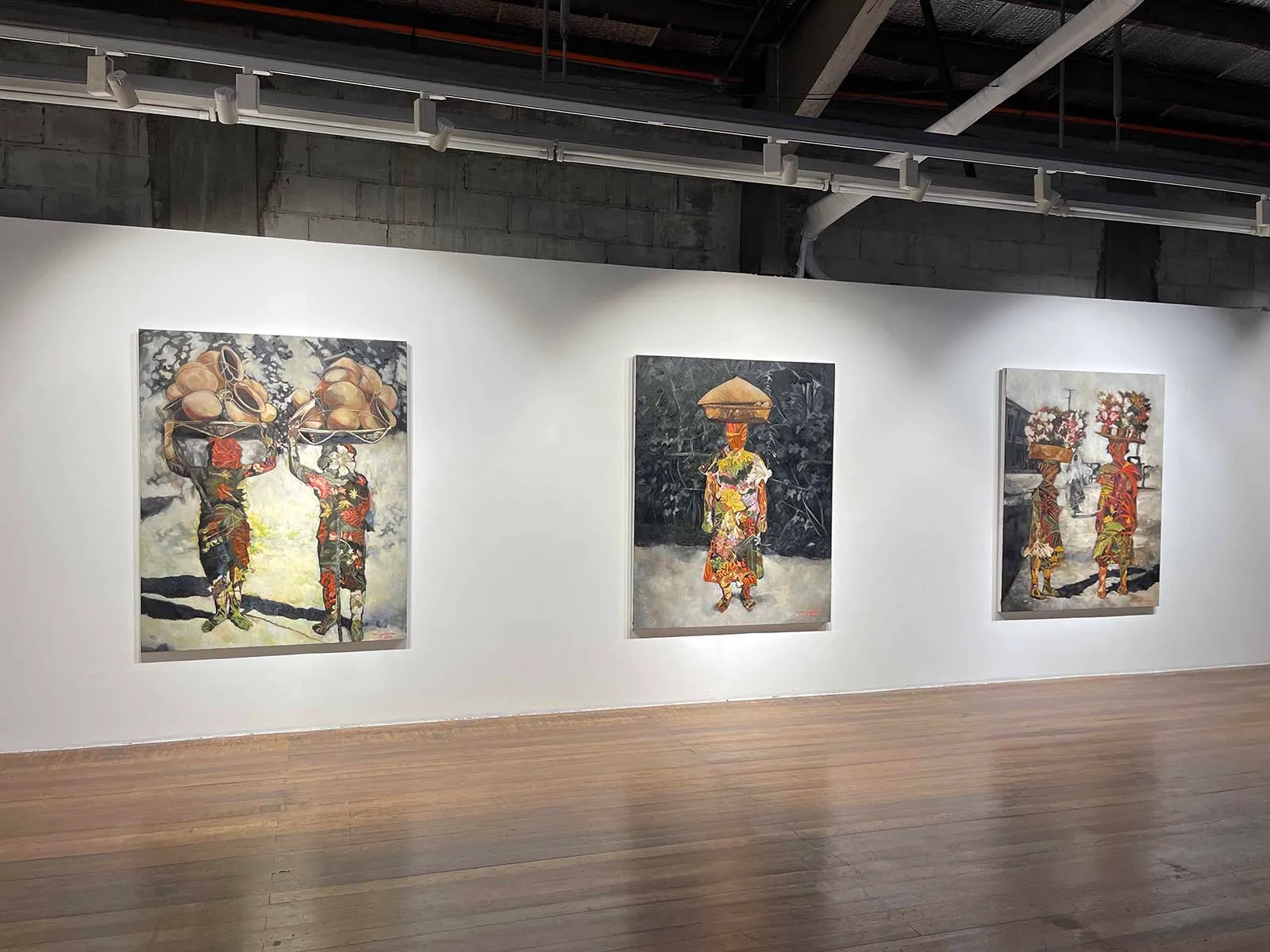 Three large paintings on a white gallery wall show figures in colorful traditional clothing carrying baskets on their heads, set against forest and street backgrounds.