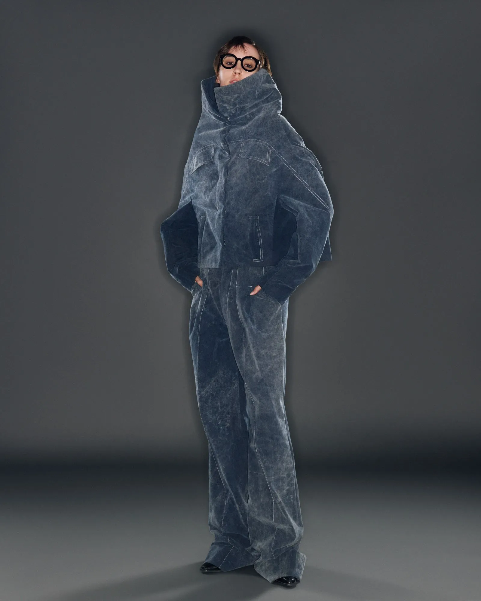 Person wearing an oversized blue denim jacket and large round black glasses stands against a dark grey background.