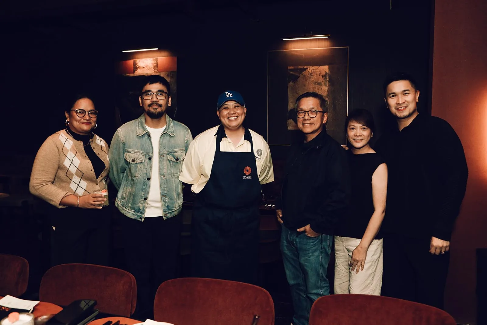 Six people stand side by side in a dimly lit restaurant, with a chef in an apron and LA cap in the center.