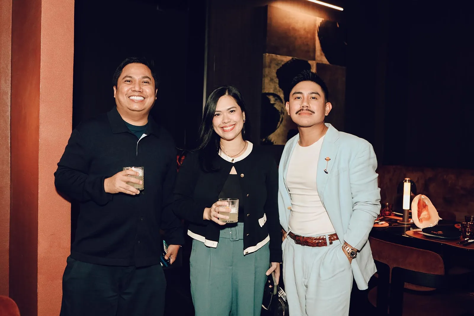 Three friends posing together at a dimly lit venue, each holding a drink and smiling at the camera.