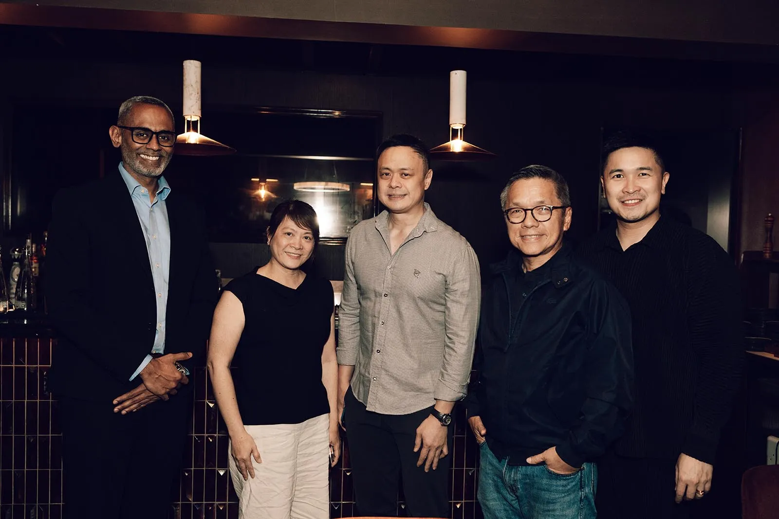 Group of five adults posing together in a dimly lit bar or restaurant, smiling at the camera.