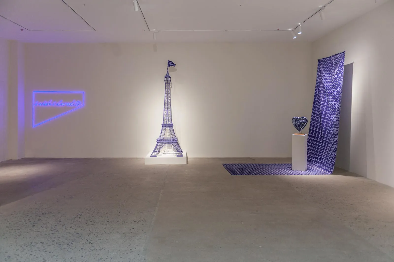 White-walled gallery with a lit blue Eiffel Tower sculpture on a pedestal in the center and a pink-purple neon sign on the left wall.