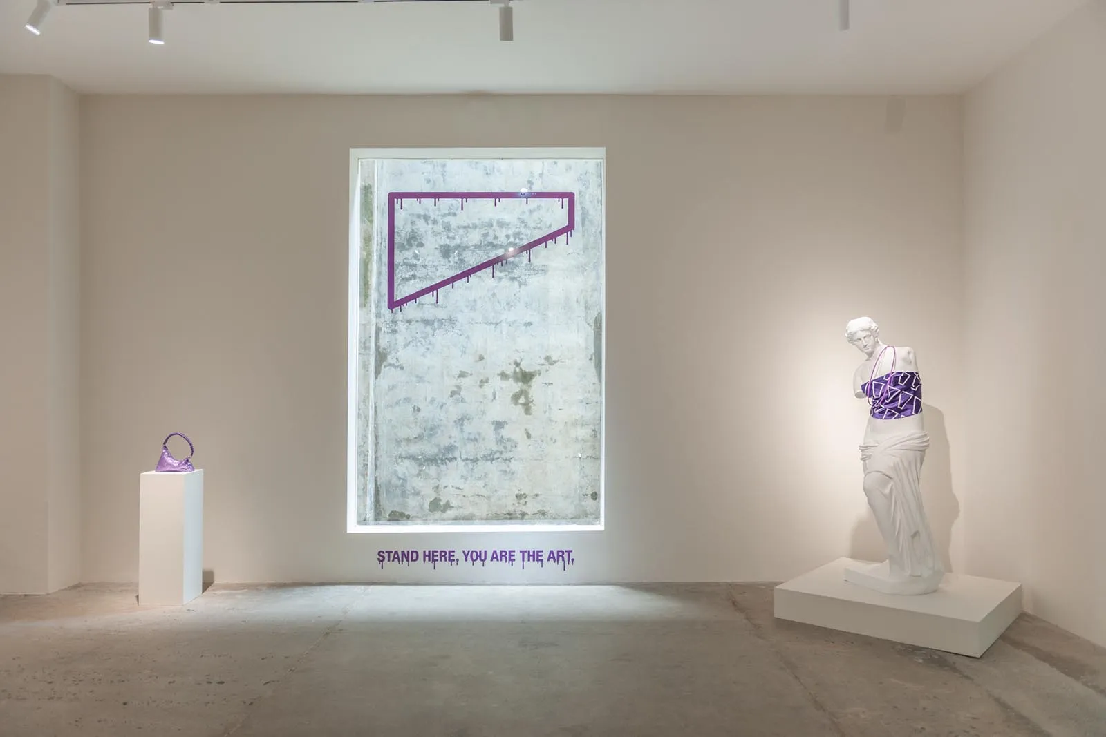 Gallery scene with a small purple purse on a pedestal left, a white statue wrapped in purple fabric on the right, and a framed concrete panel featuring a purple geometric outline; text underneath reads 'STAND HERE, YOU ARE THE ART'.